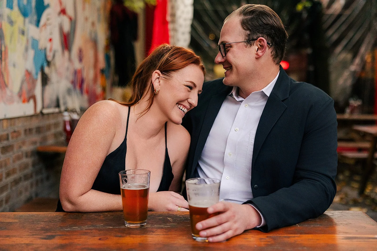 A couple sitting closely together at a bar or restaurant, sharing a moment of laughter. The woman has red hair and is wearing a black spaghetti strap dress, holding a glass of beer. The man has dark hair, glasses, and is dressed in a dark blazer over