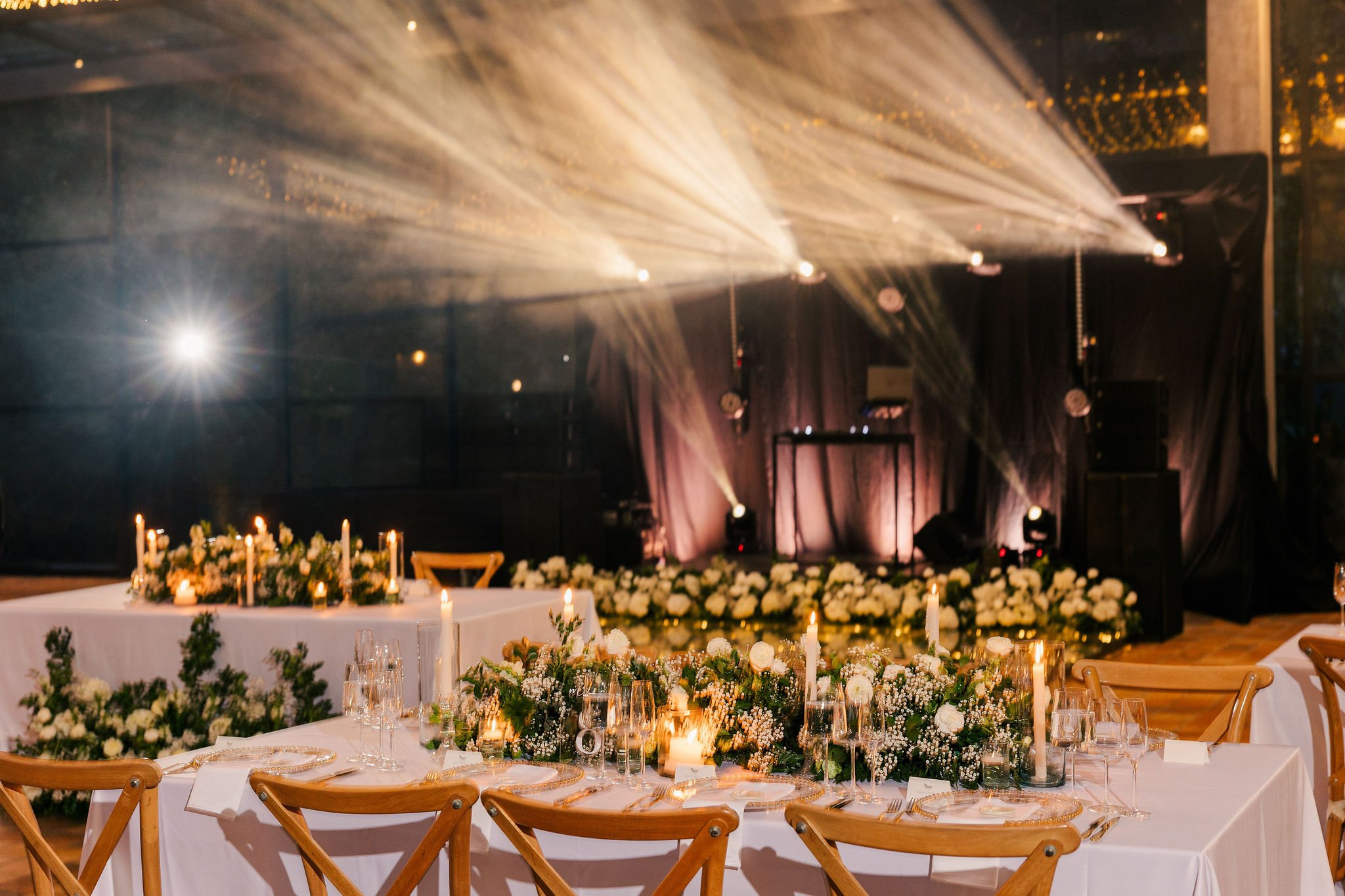 Elegant event setup with a long dining table decorated with candles and floral arrangements, a stage with lighting and sound equipment in the background, and warm lighting creating a cozy atmosphere.