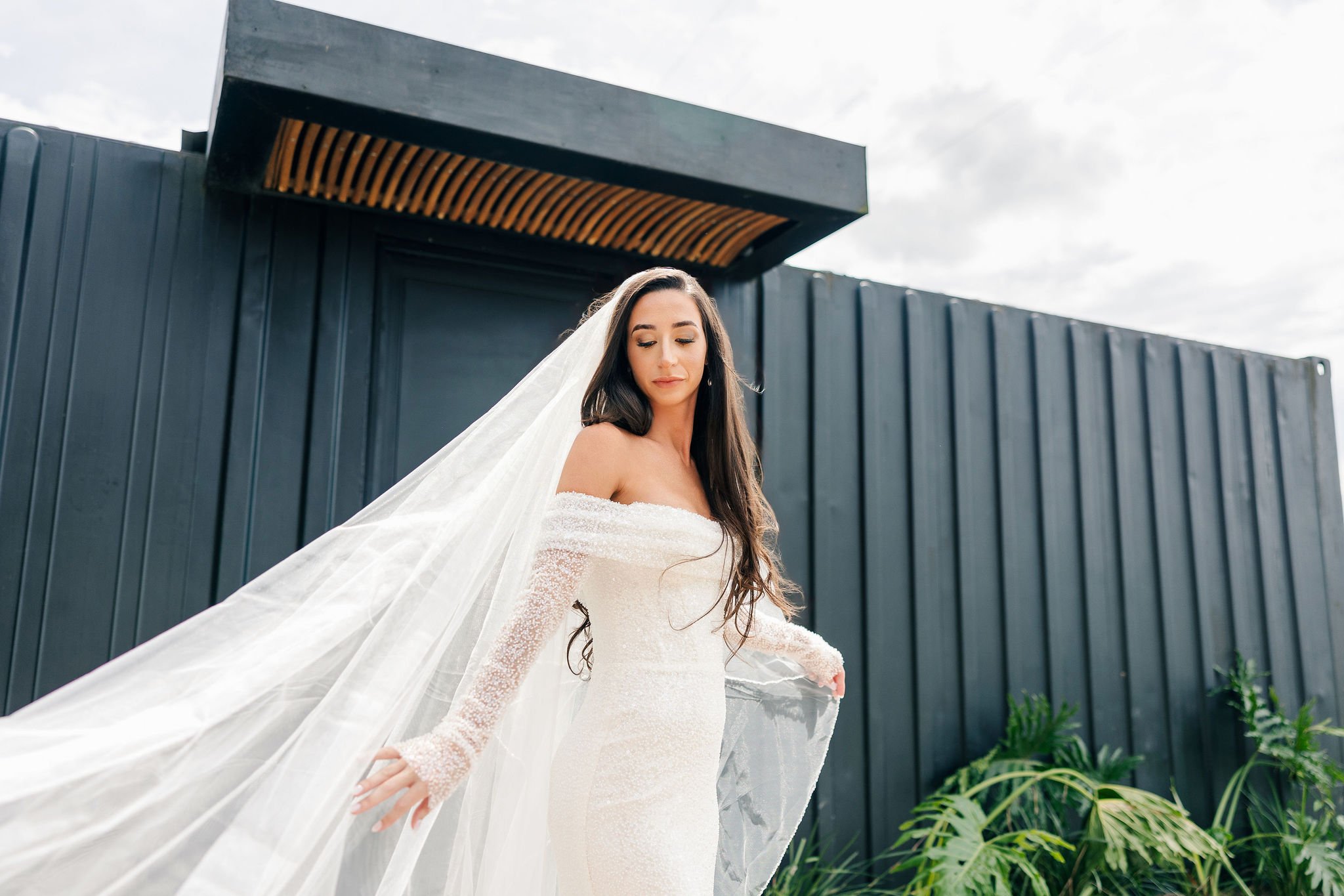 A bride with long dark hair wearing an off-the-shoulder white wedding gown with a long veil standing outdoors in front of a modern dark gray building with plants at her side.