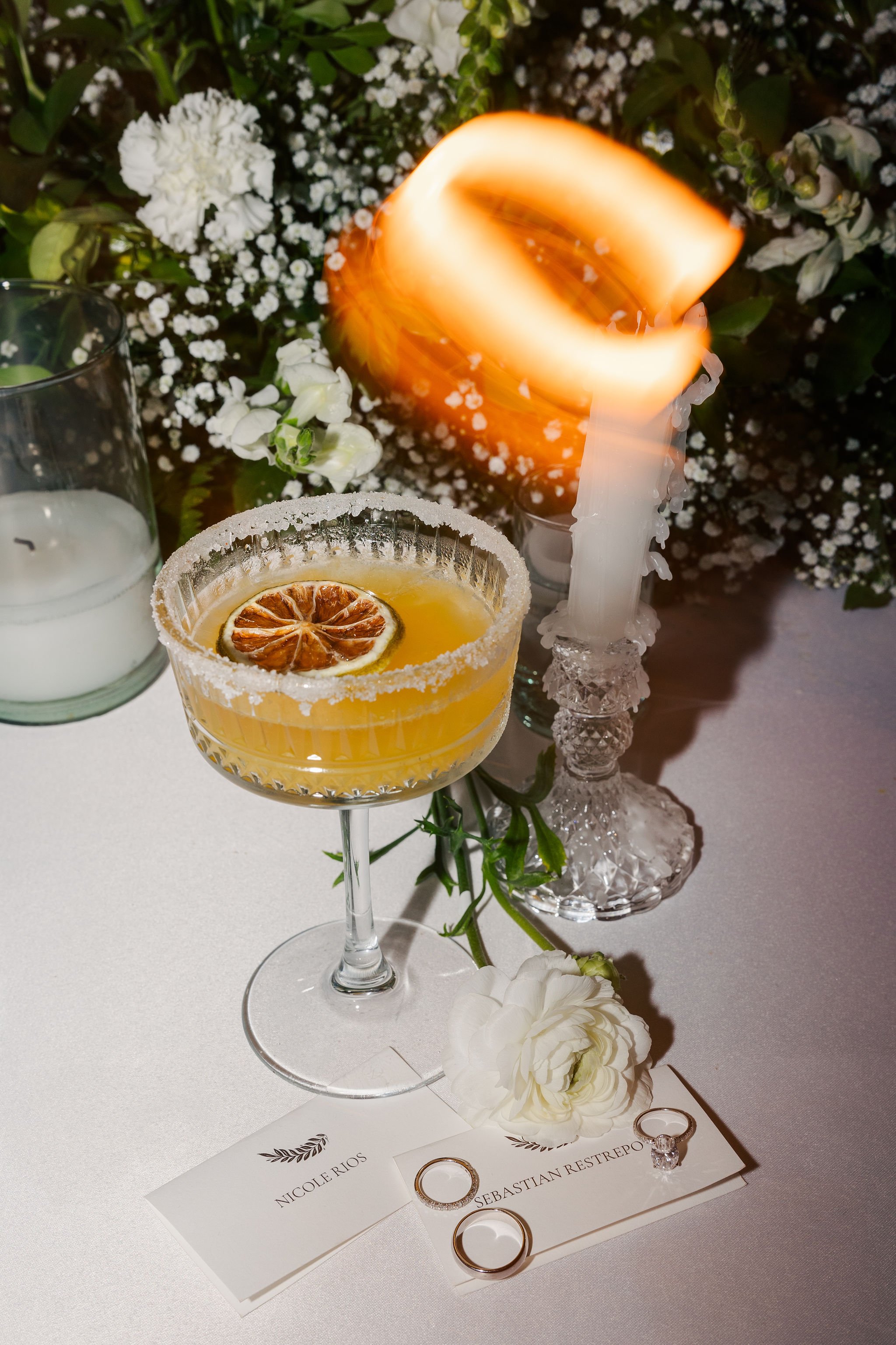 A festive table setting with a lemon cocktail garnished with a dried lemon slice, surrounded by white flowers, candles, and wedding rings on place cards, at an elegant event or wedding reception.