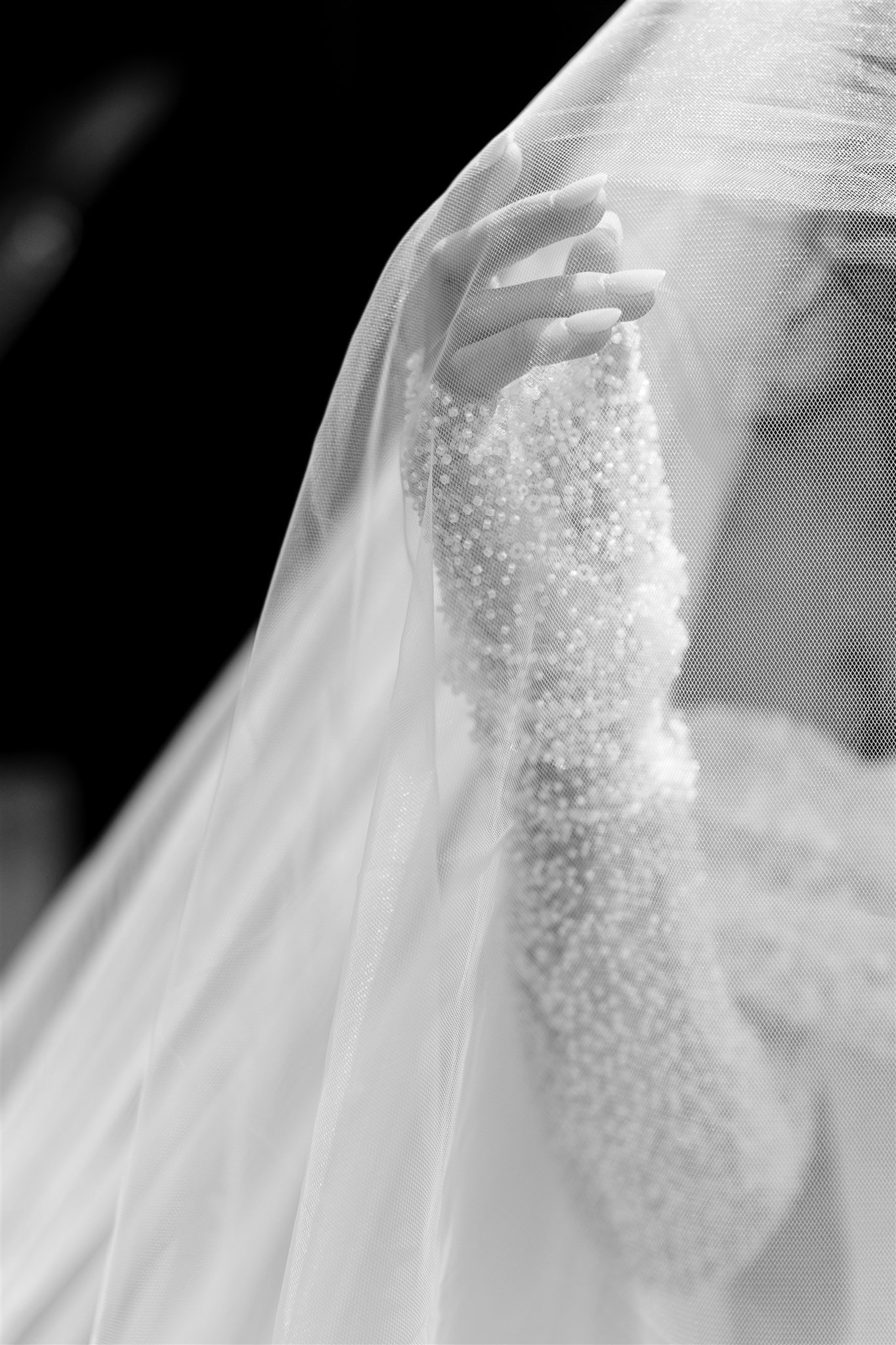 Close-up black and white photo of a woman's hand holding a veil over her shoulder, with a beaded or pearl embellishment on her dress, set against a dark background.