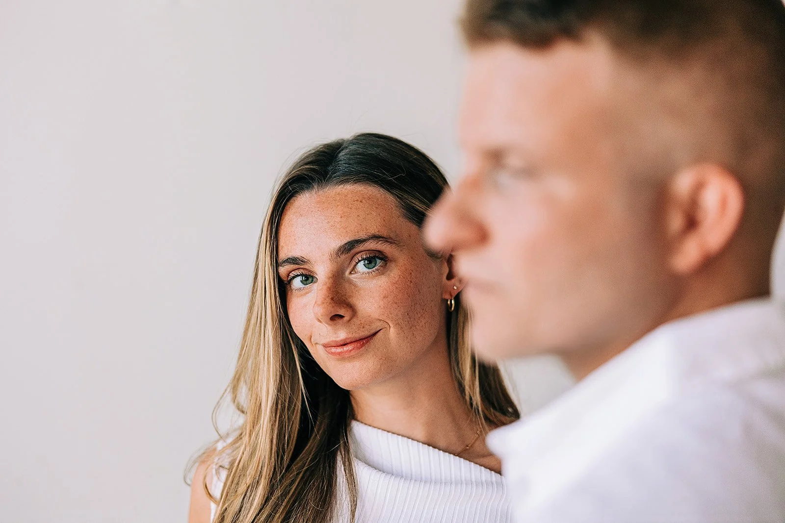 A woman with long dark hair and blue eyes smiling, looking at a man with short hair and glasses, who is slightly out of focus and facing sideways.