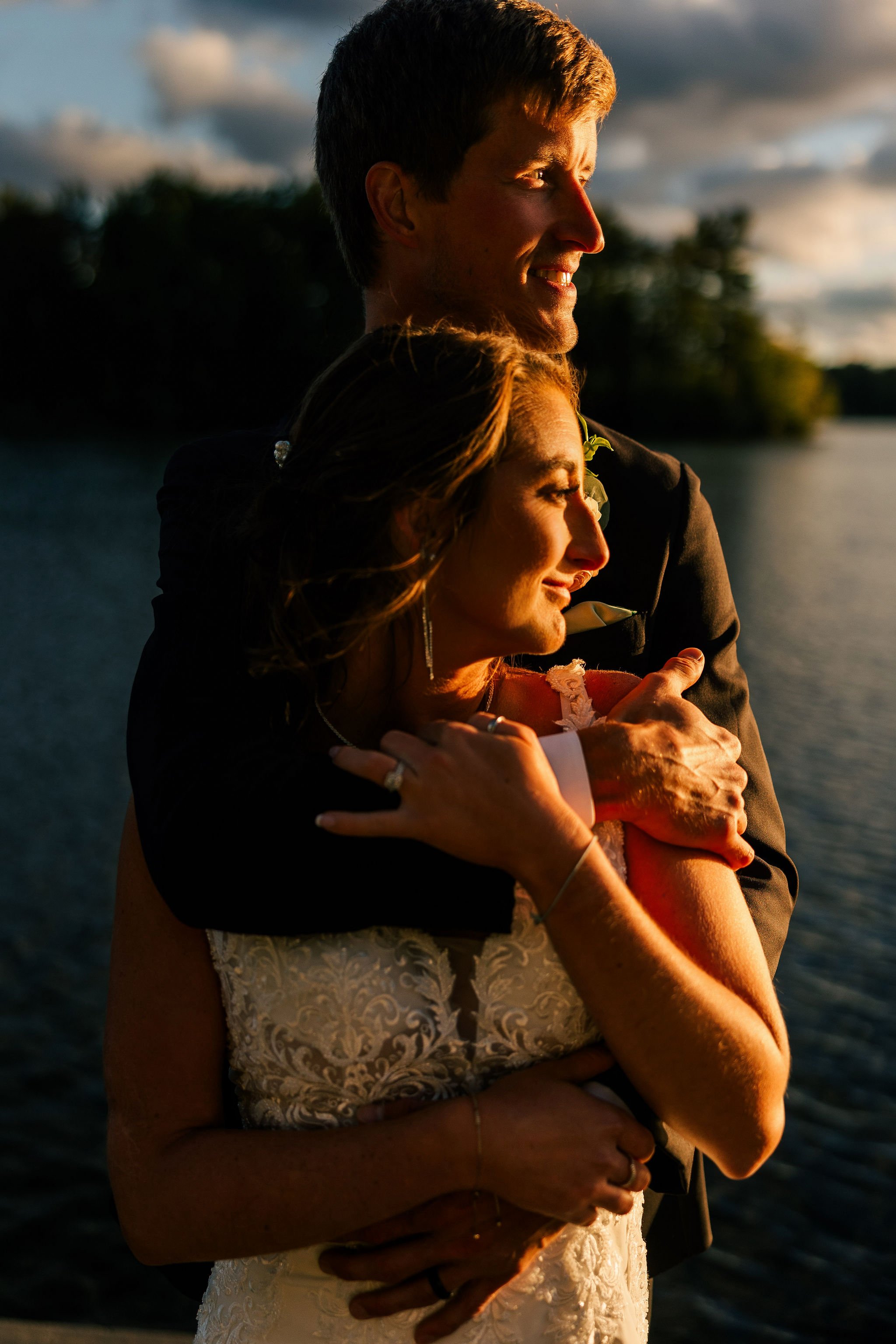 A newlywed couple embracing near a body of water at sunset, with the man hugging the woman from behind, both smiling softly.