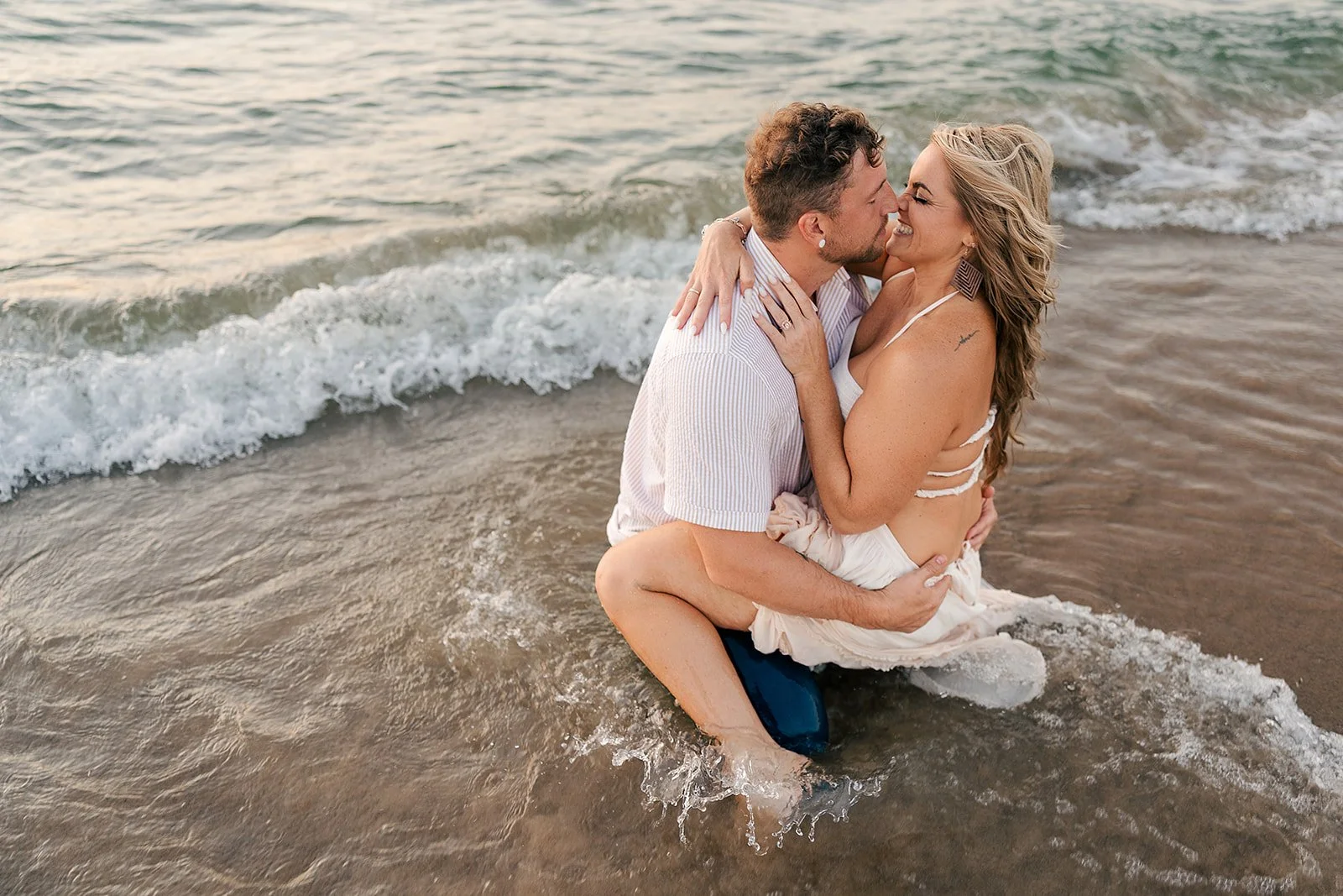 A couple sharing a kiss while sitting in the shallow water at the beach during sunset.