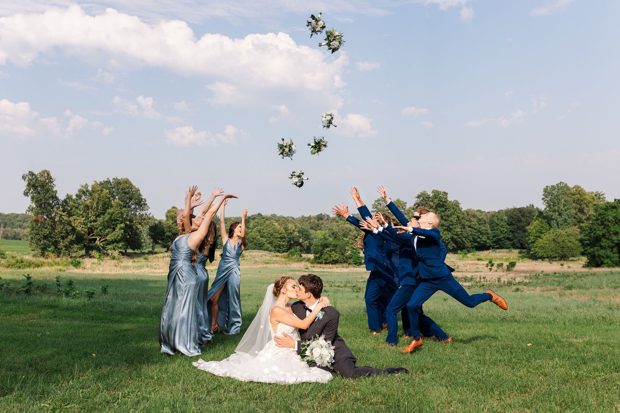 Bride and groom sitting on grass, kissing, surrounded by wedding party throwing bouquets and jumping in a green field with trees and blue sky.