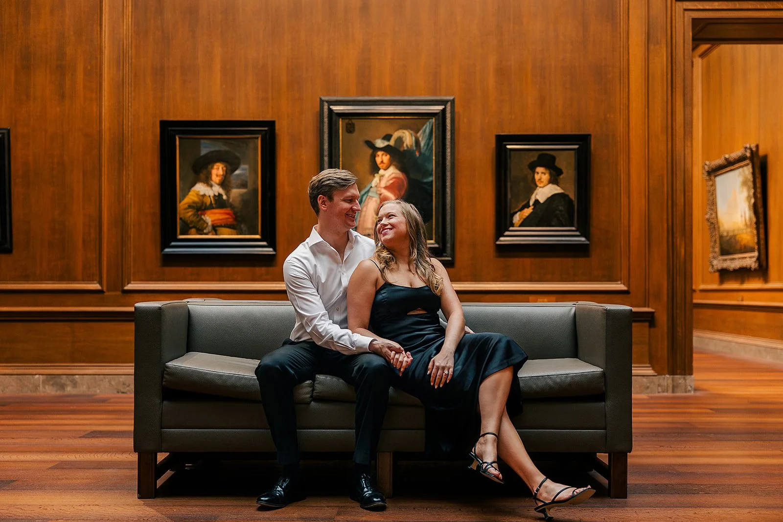 A couple sitting on a gray sofa in an art gallery, smiling at each other. The woman is wearing a black dress and heels, and the man is in a white shirt and black pants. Behind them are wooden-paneled walls with framed classical portraits.