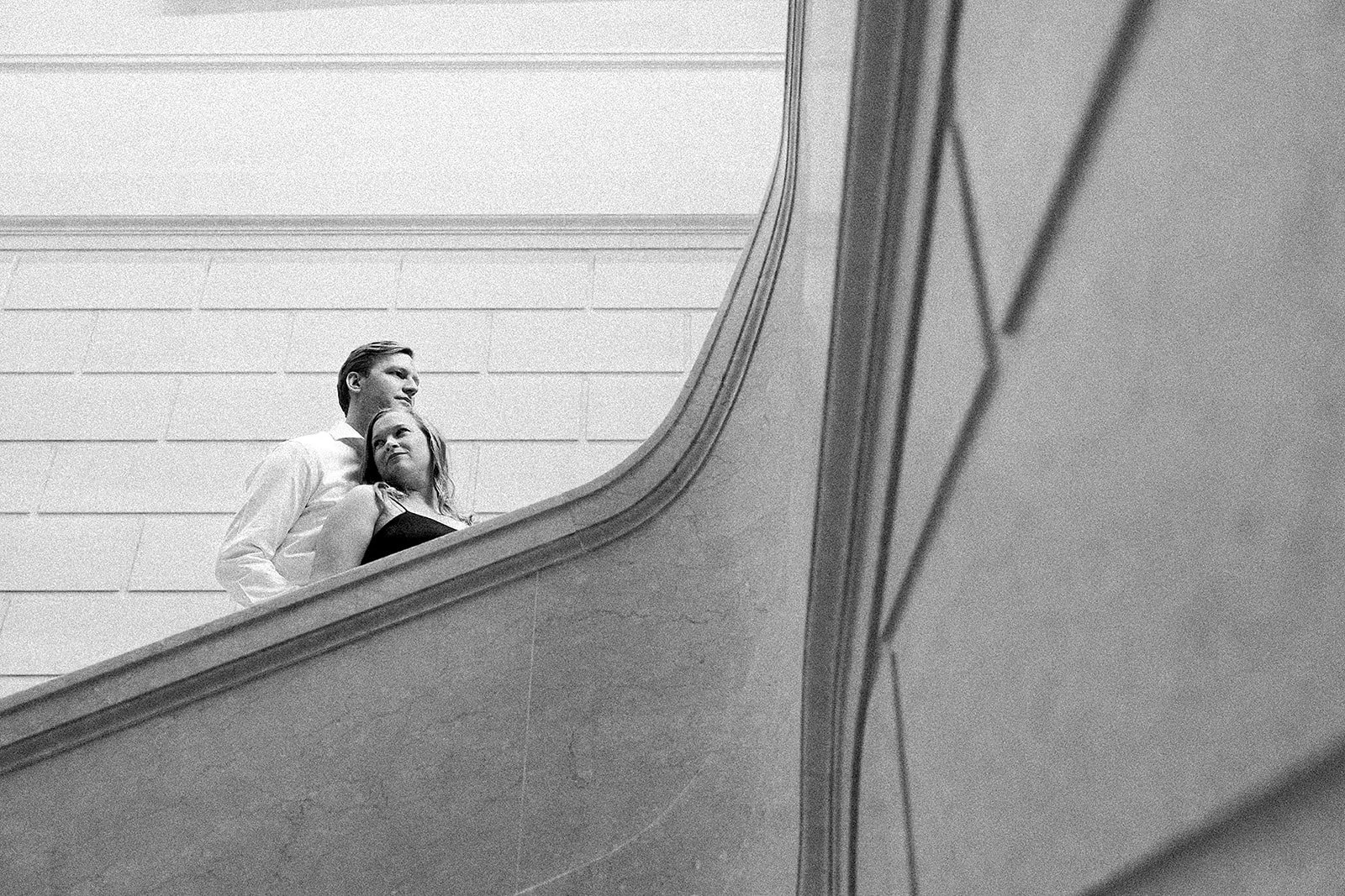 Two people, a man and a woman, standing on a staircase and looking outward. The photo is in black and white.