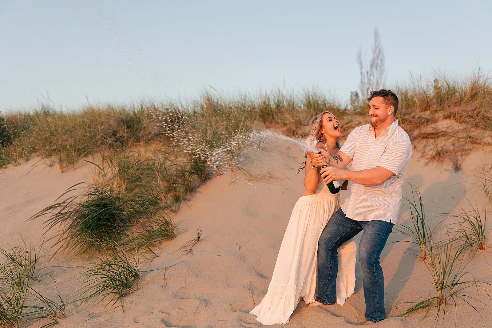 A couple celebrating on a sandy beach, spraying champagne together and smiling.