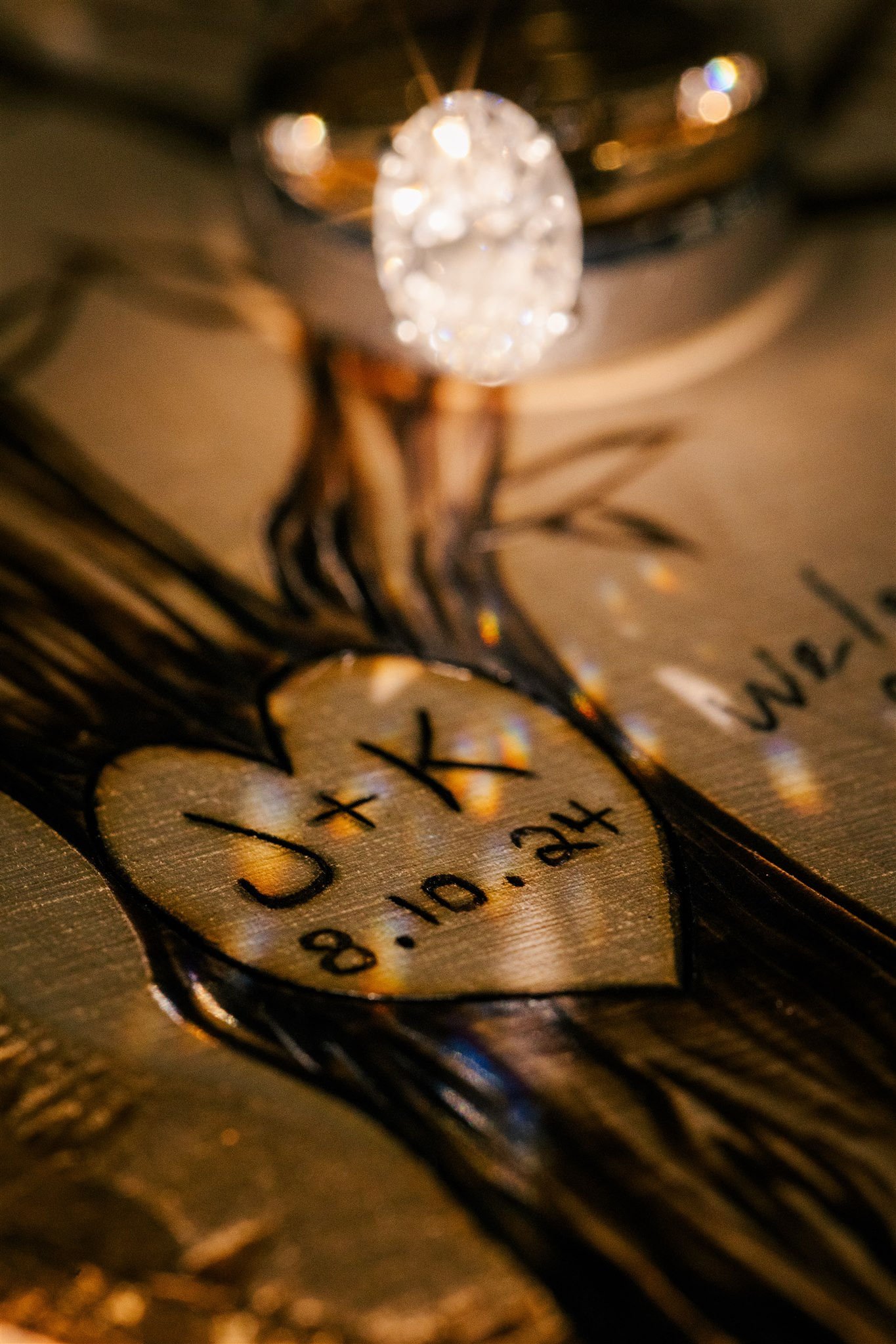 Close-up of a textured metal surface with a heart-shaped engraving containing initials and a date, and a glass ornament above. The engraving reads J + T 8.10.24.