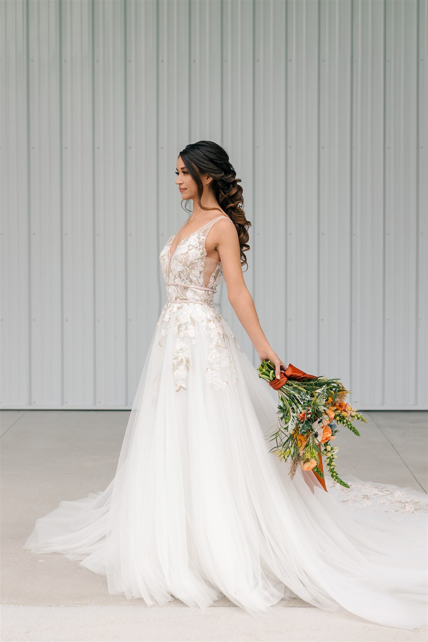 Bride in white lace wedding gown holding a bouquet of flowers, standing against a light gray metal wall.