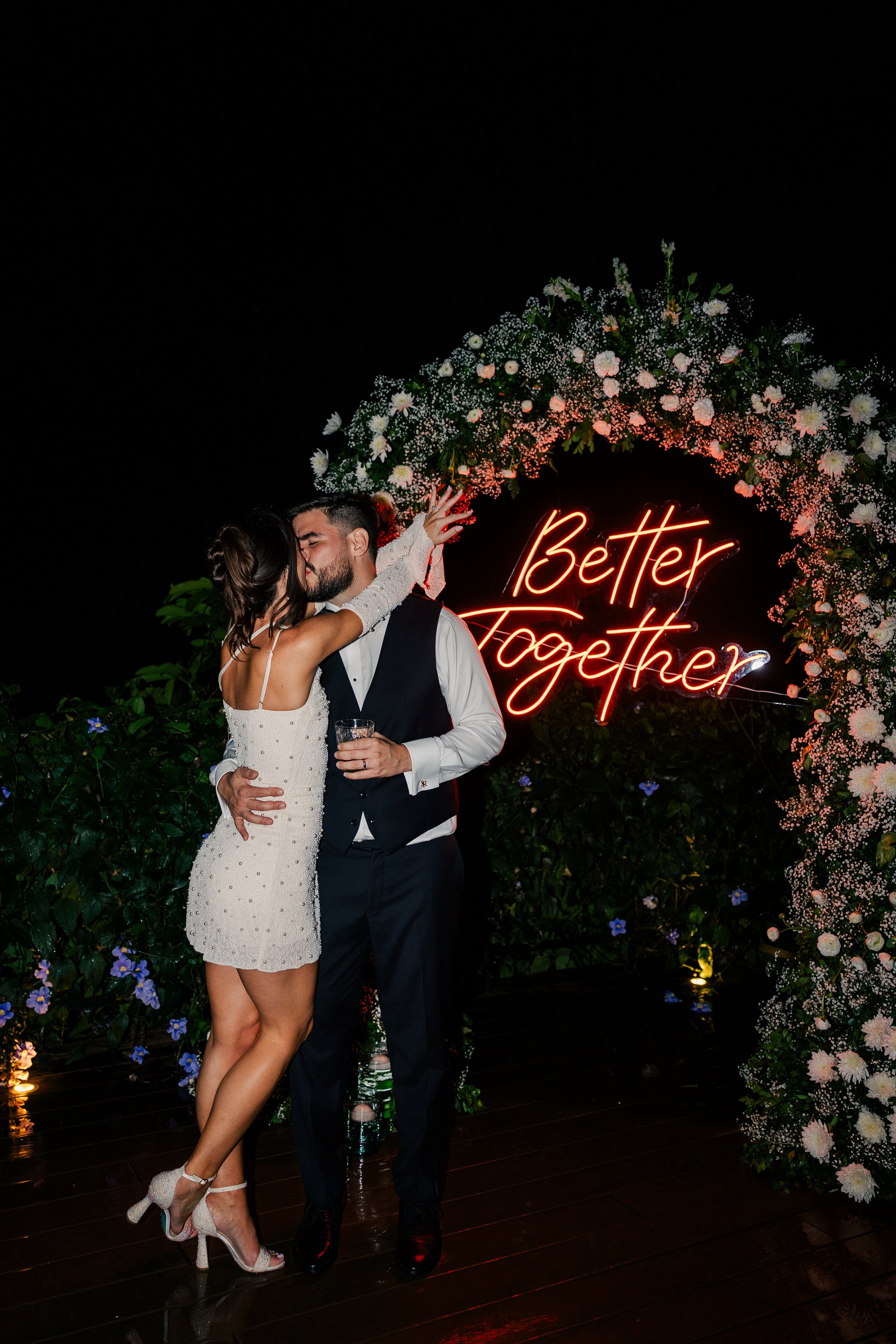 A couple kissing at night in front of a neon sign that says 'Better Together' surrounded by flowers.
