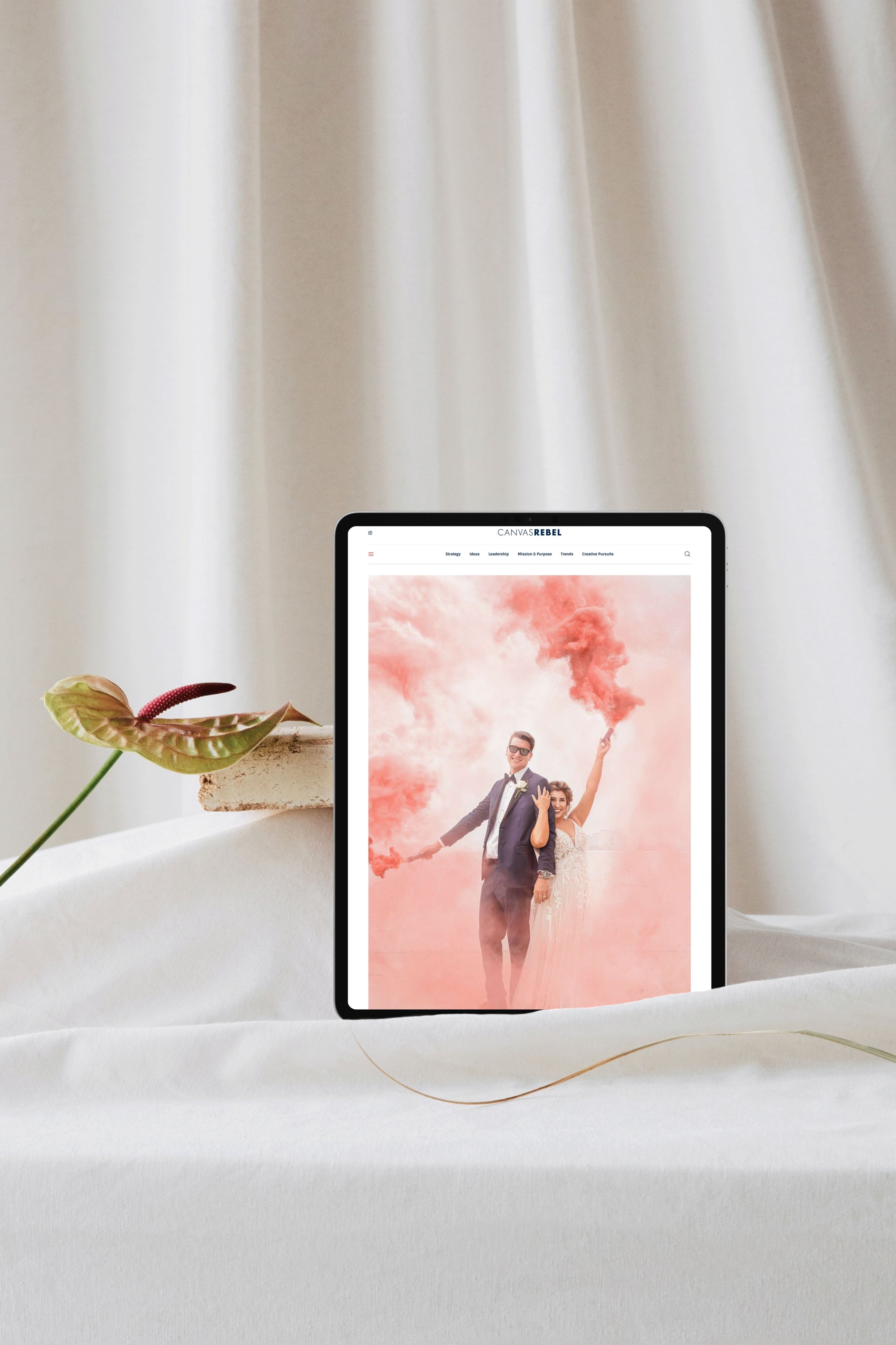 Digital tablet displaying a wedding photo of a couple in formal attire, standing among pink smoke clouds, with a plant and fabric backdrop.