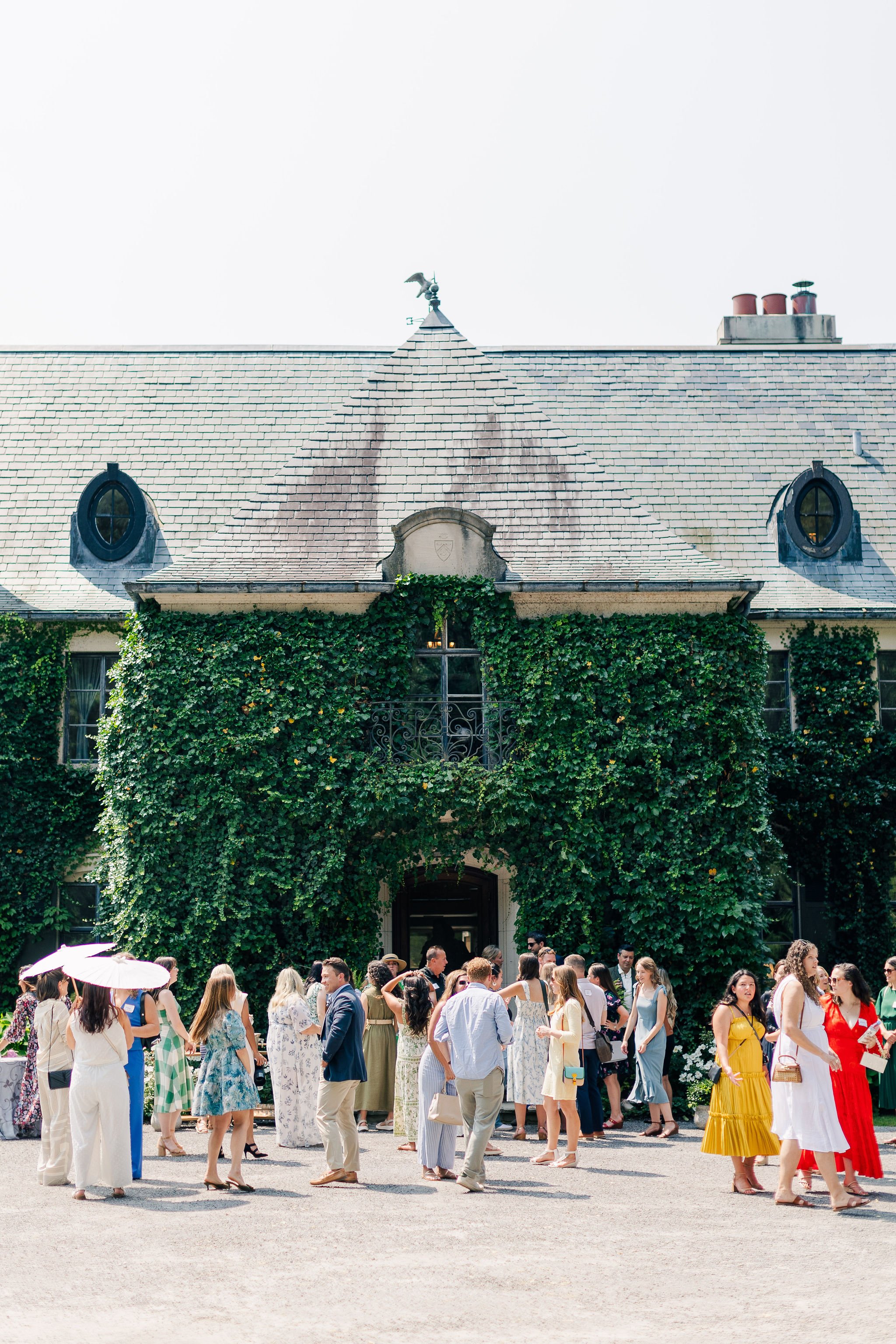 A group of people gathered outside a mansion with ivy-covered walls and a slate roof during daytime.