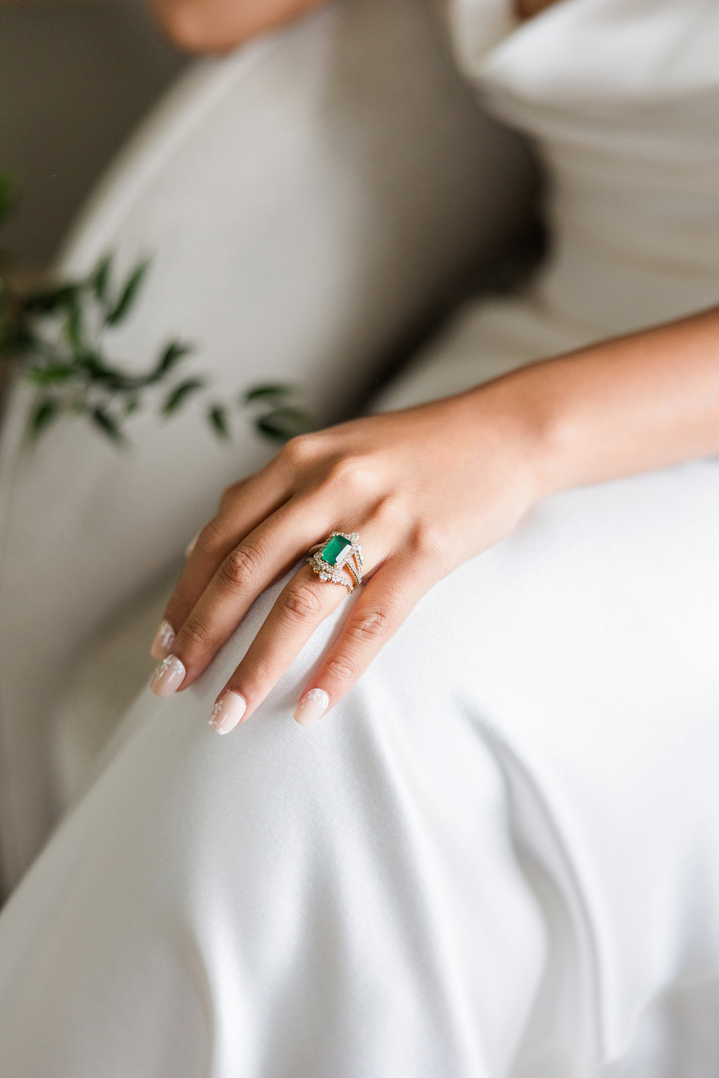 Close-up of a woman's hand resting on her lap, showcasing an emerald and diamond ring with intricate gold detailing, against white satin fabric.