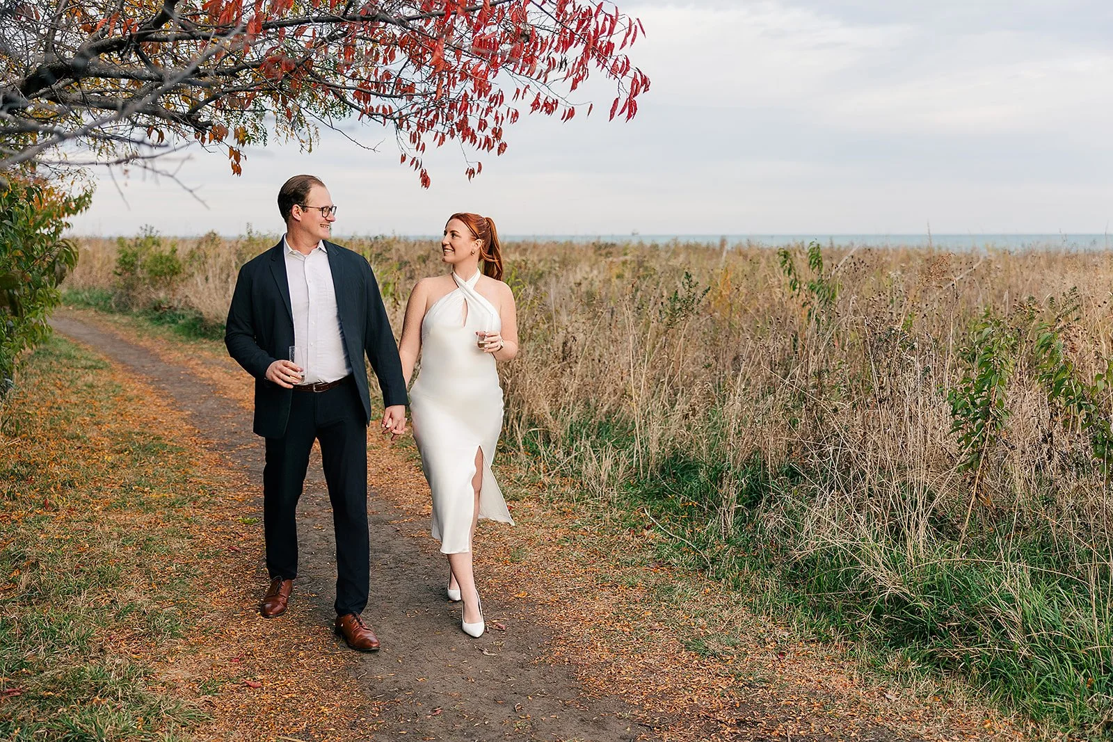 A couple walking hand in hand on a dirt path through a grassy field during fall, holding drinks and smiling at each other under a partly cloudy sky.