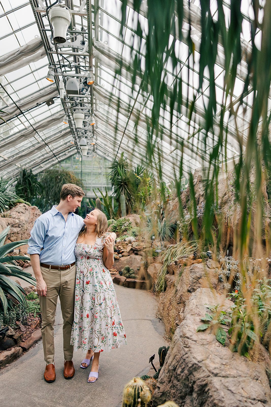 A couple walking inside a lush tropical greenhouse, holding hands, and looking into each other's eyes, surrounded by various green plants and cacti.