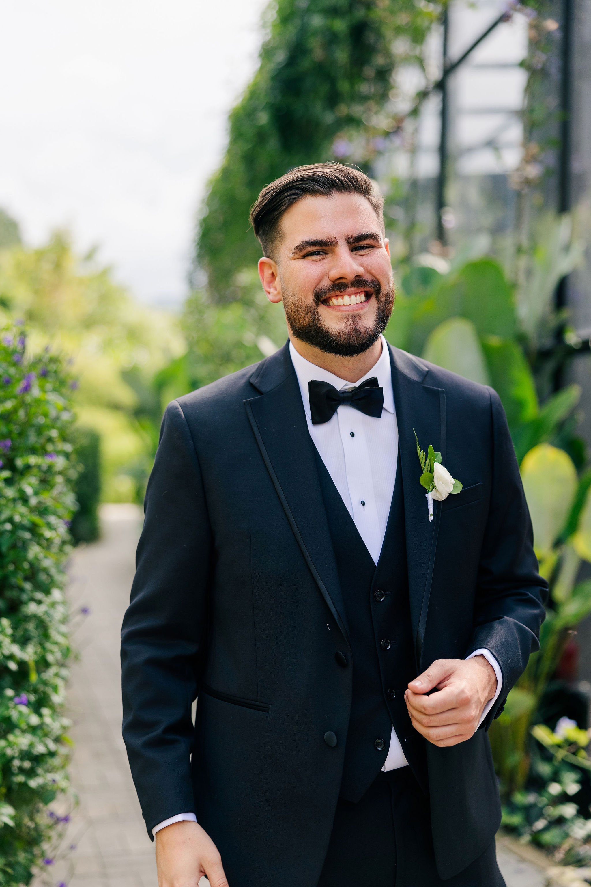 A smiling man in a black tuxedo with a boutonnière, standing outdoors on a pathway surrounded by greenery and plants.