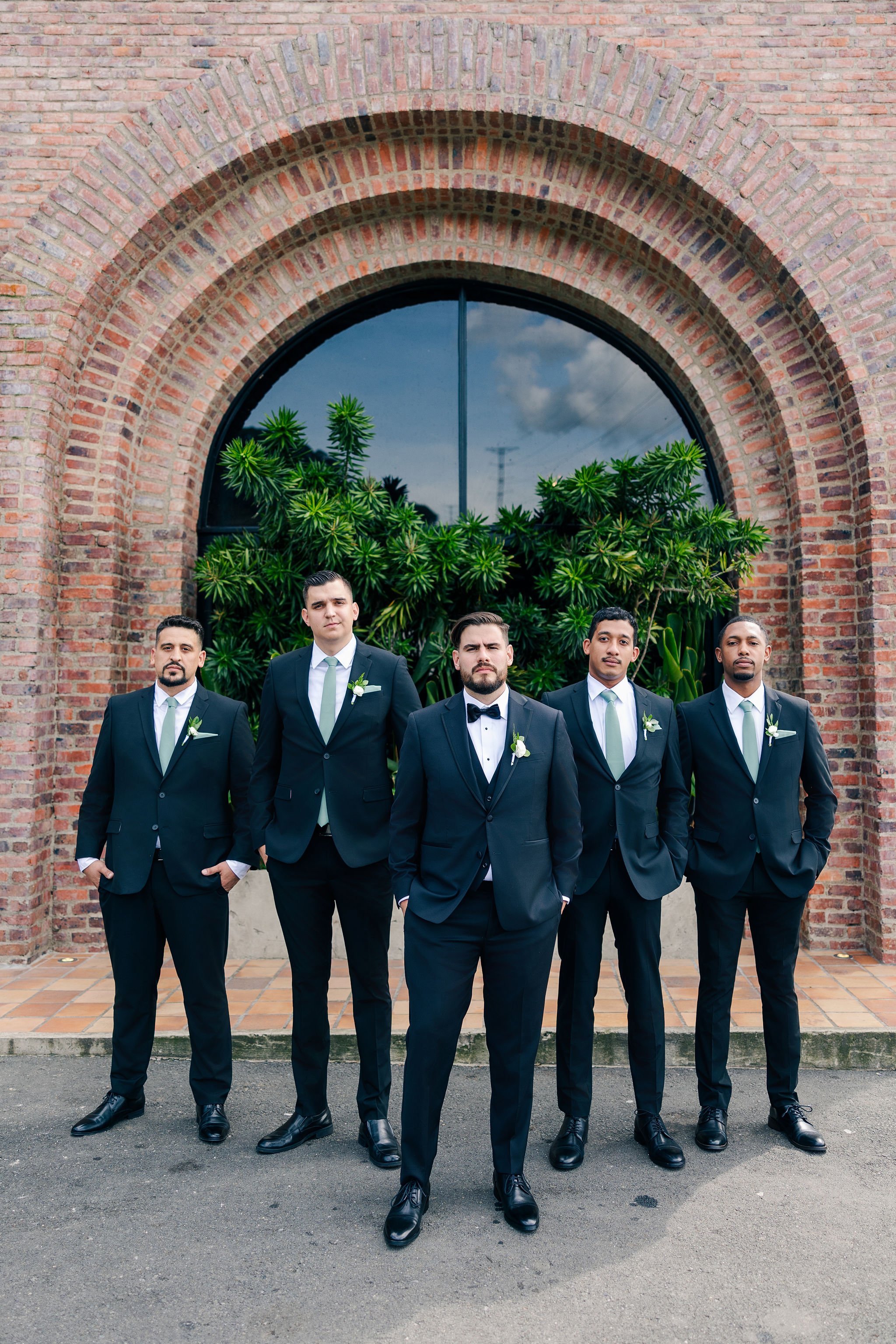 Group of five men dressed in formal suits, standing in front of a brick building with a large window and greenery.