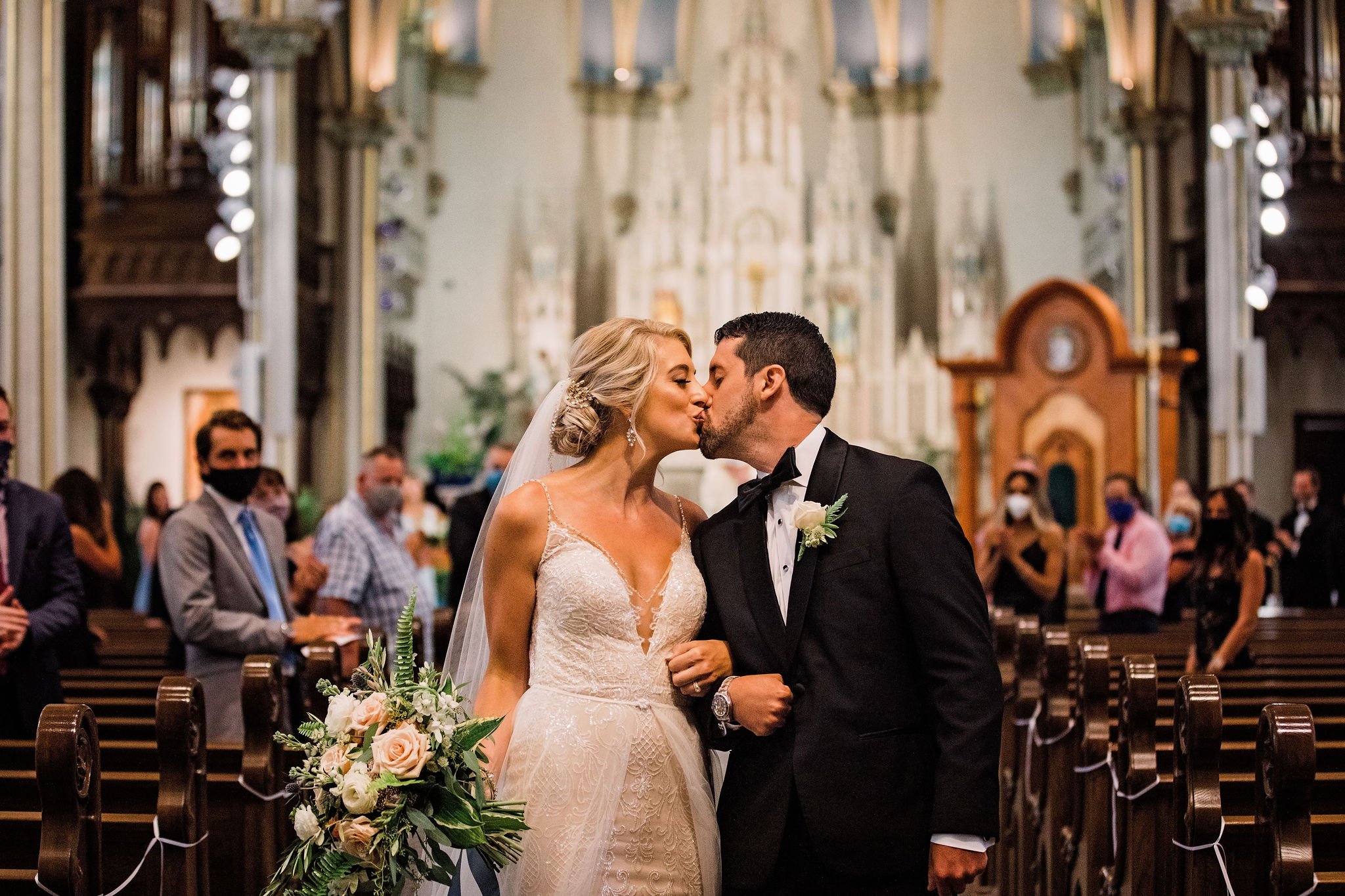 A bride and groom kiss inside a church during their wedding ceremony, with guests watching in the background.