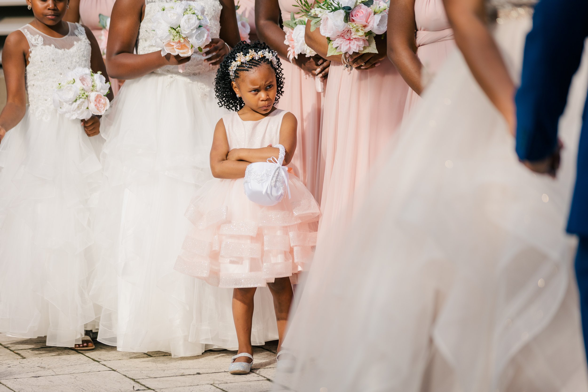 Young girl in a pink dress with arms crossed, standing among women and girls in formal dresses holding bouquets at a wedding ceremony.