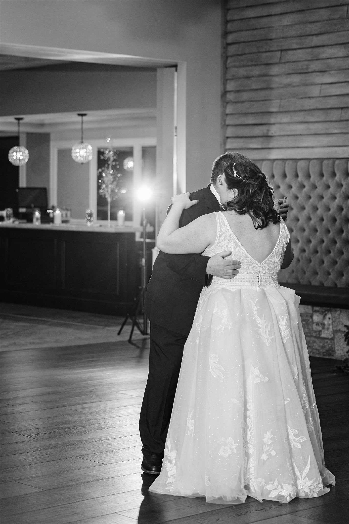 A bride and groom sharing a dance at their wedding reception, with the bride wearing a lace wedding gown and the groom in a dark suit, illuminated by a bright light behind them.