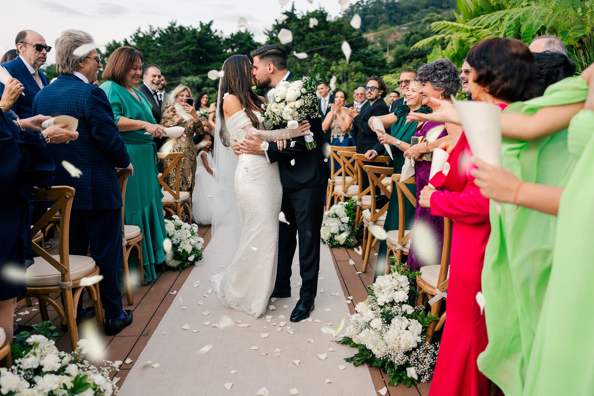 A newlywed couple sharing a kiss during their outdoor wedding ceremony, surrounded by their guests throwing flower petals.
