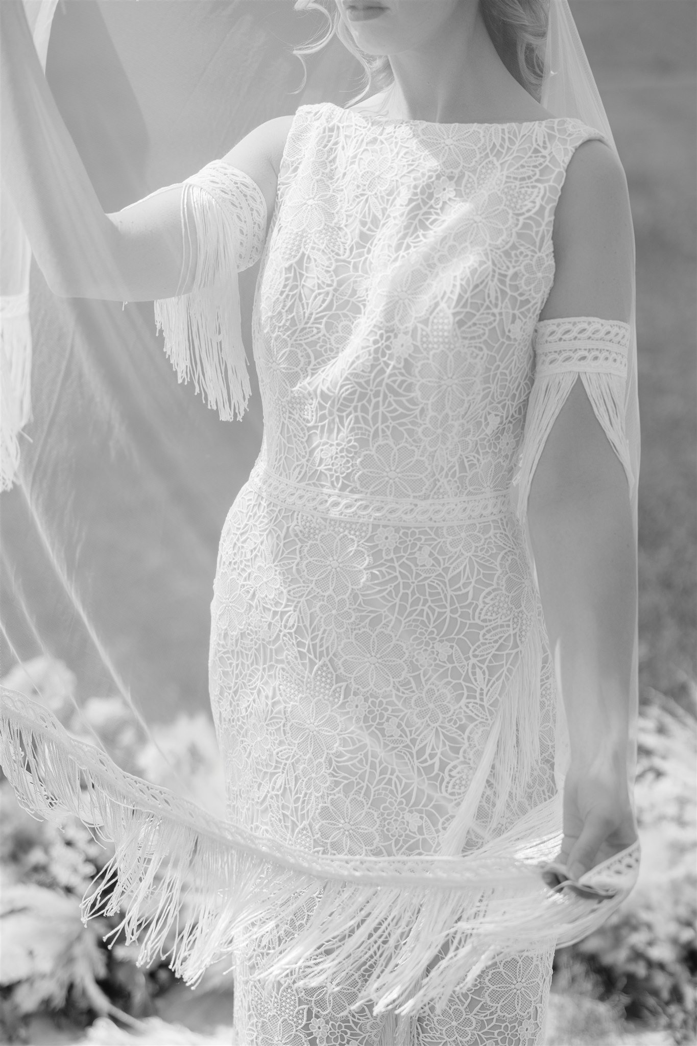 A woman wearing a lace wedding dress with cut-out shoulders, holding a fringed fabric in a field.