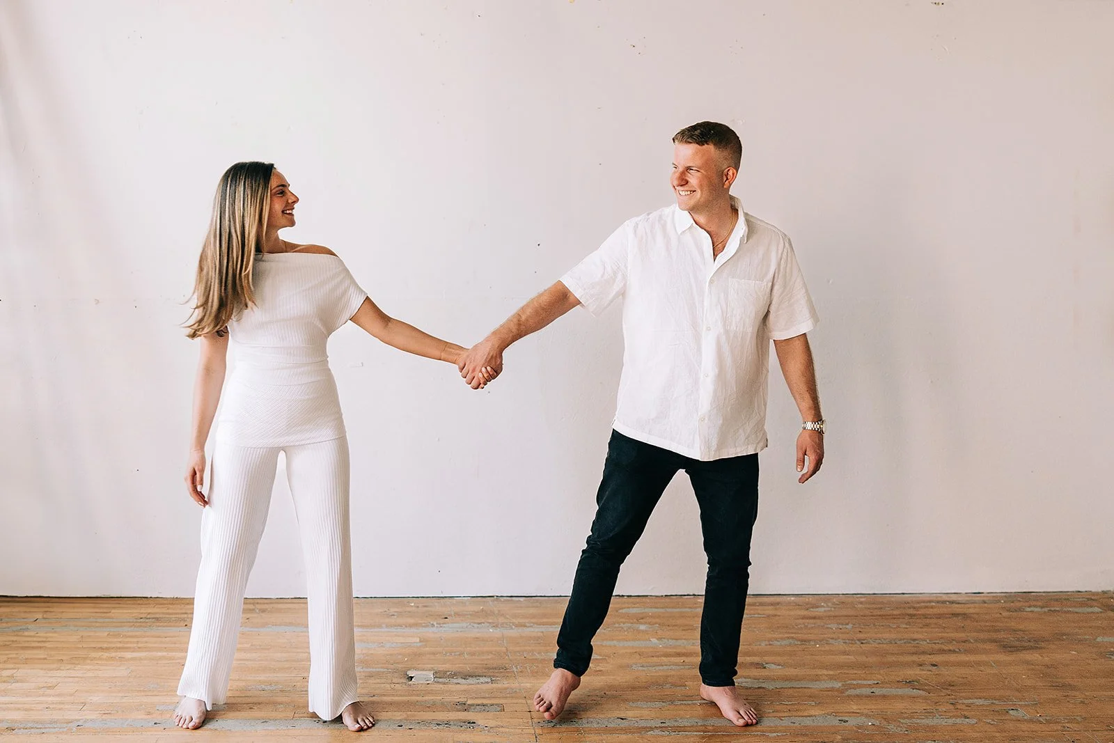 A man and woman holding hands and smiling, standing on a wooden floor against a plain white wall.