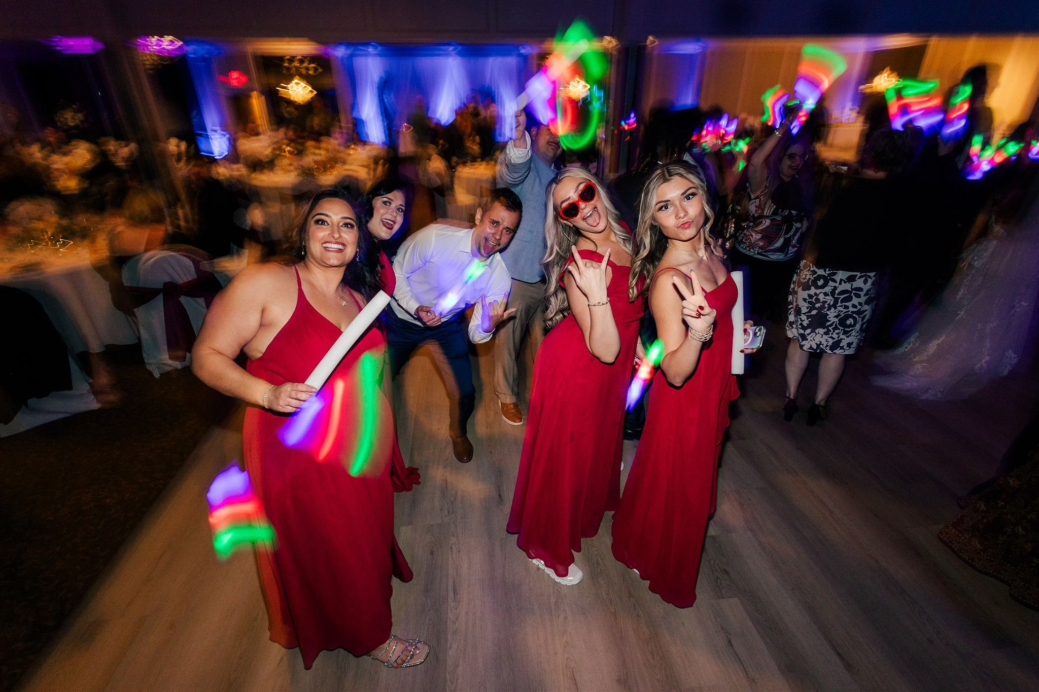 Group of five people dancing and posing at a party, with colorful lights and blurred background, four women in red dresses and one man in white shirt.
