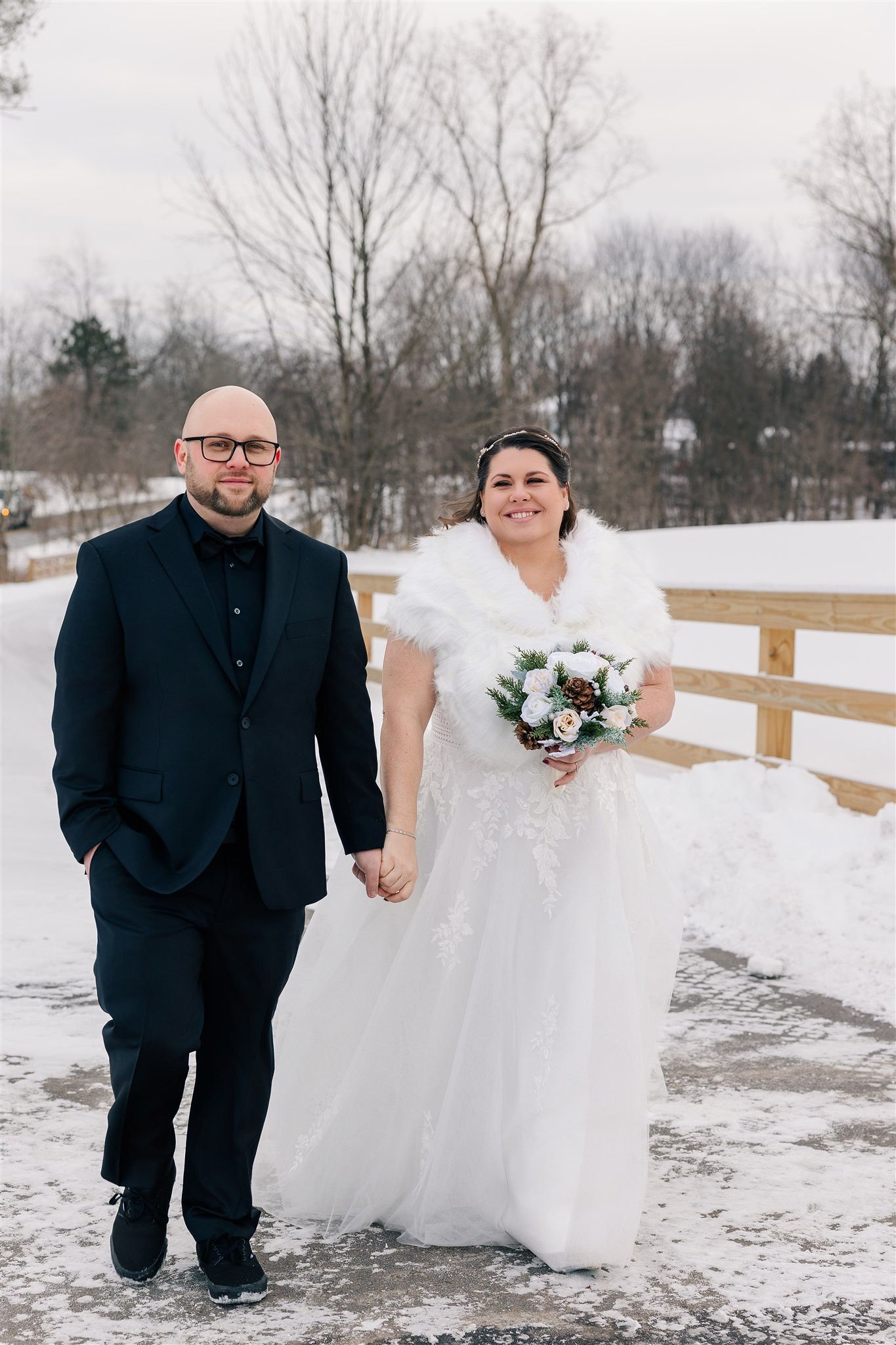 A newlywed couple holding hands outdoors on a snowy day, with the bride in a white wedding dress and fur cape, and the groom in a black suit, standing on a snow-covered pathway with trees in the background.