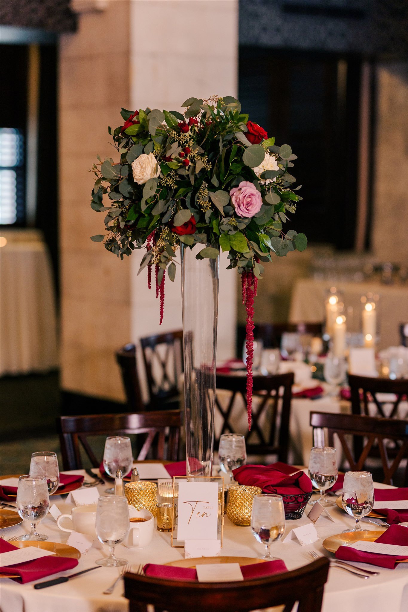 Elegant round wedding reception table decorated with a tall floral centerpiece, gold and clear candle holders, red napkins, glasses of water, and table number card in a gold frame.