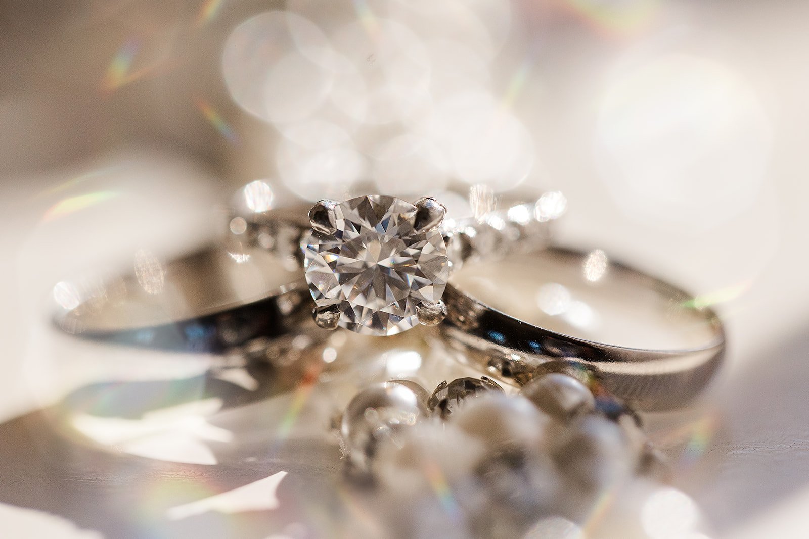 Close-up of a diamond engagement ring with a round-cut center stone, surrounded by two wedding bands and pearl beads, with sparkling light effects in the background.