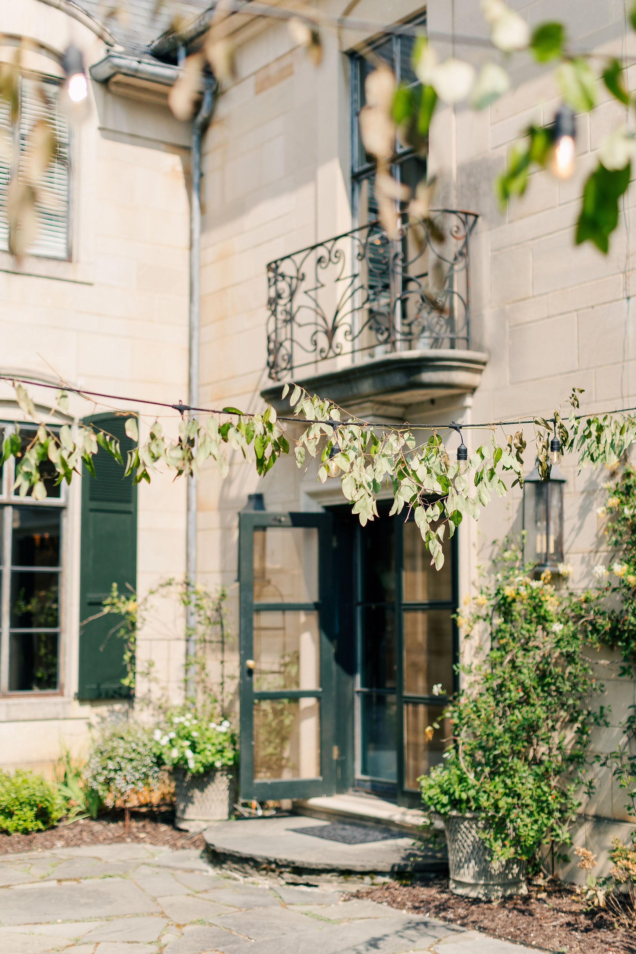 Stone house entrance with an open door, potted plants, and string lights overhead.