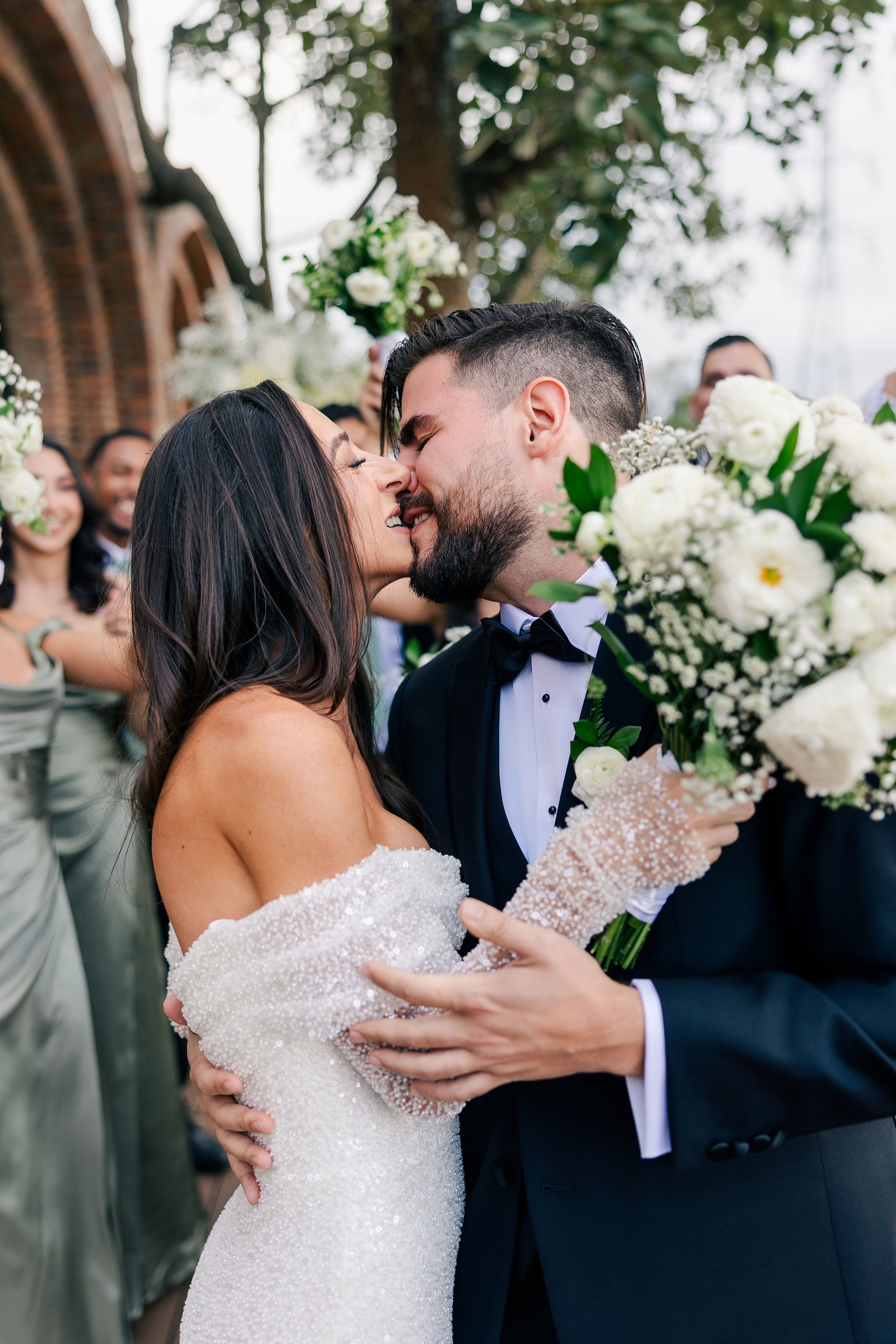 A newlywed couple about to kiss, surrounded by friends on a wedding day, with the bride holding a large bouquet of white flowers and wearing a pearl-beaded off-shoulder wedding dress.
