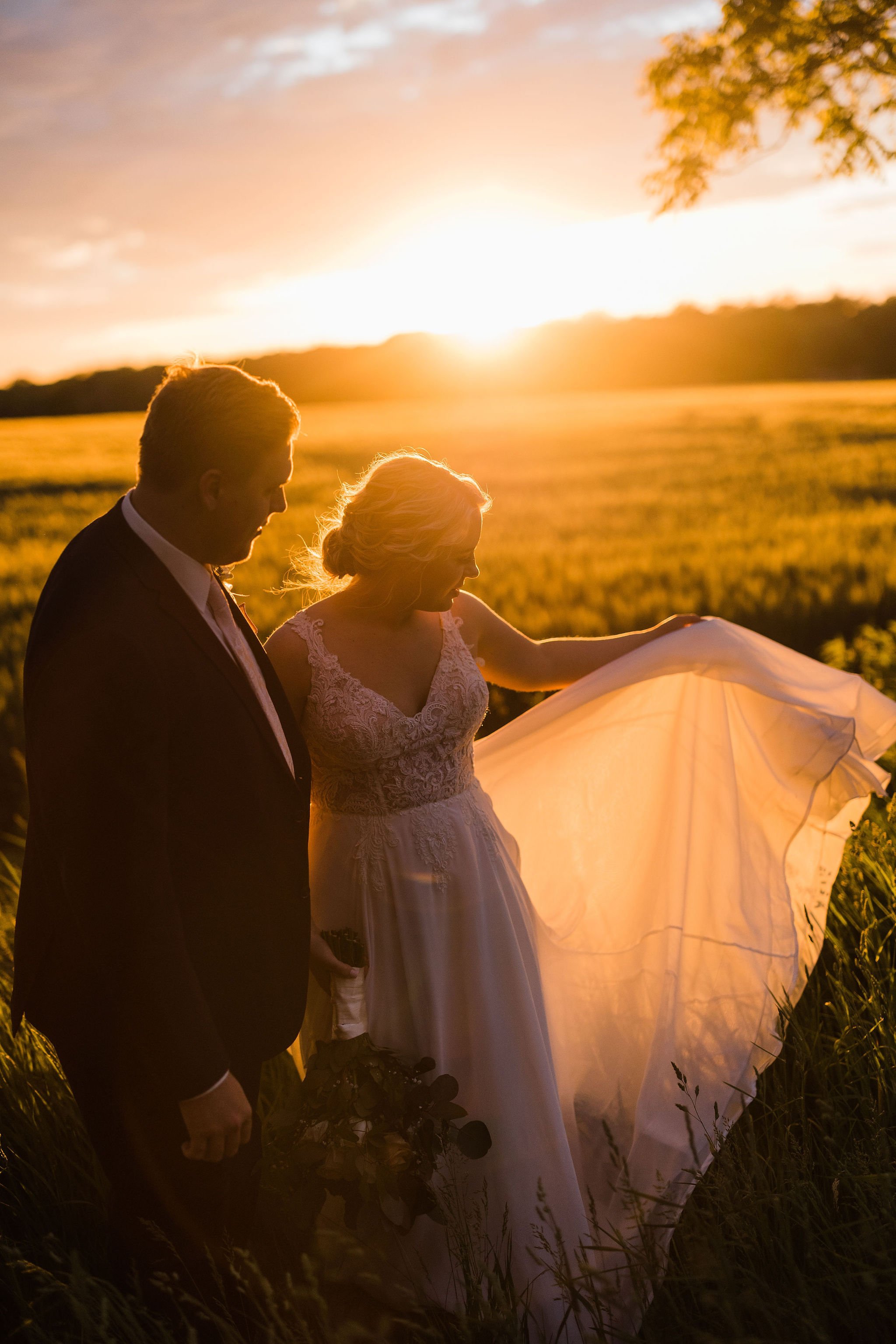 A bride and groom standing in a field at sunset, with the bride holding out her dress and the groom holding a bouquet of flowers.