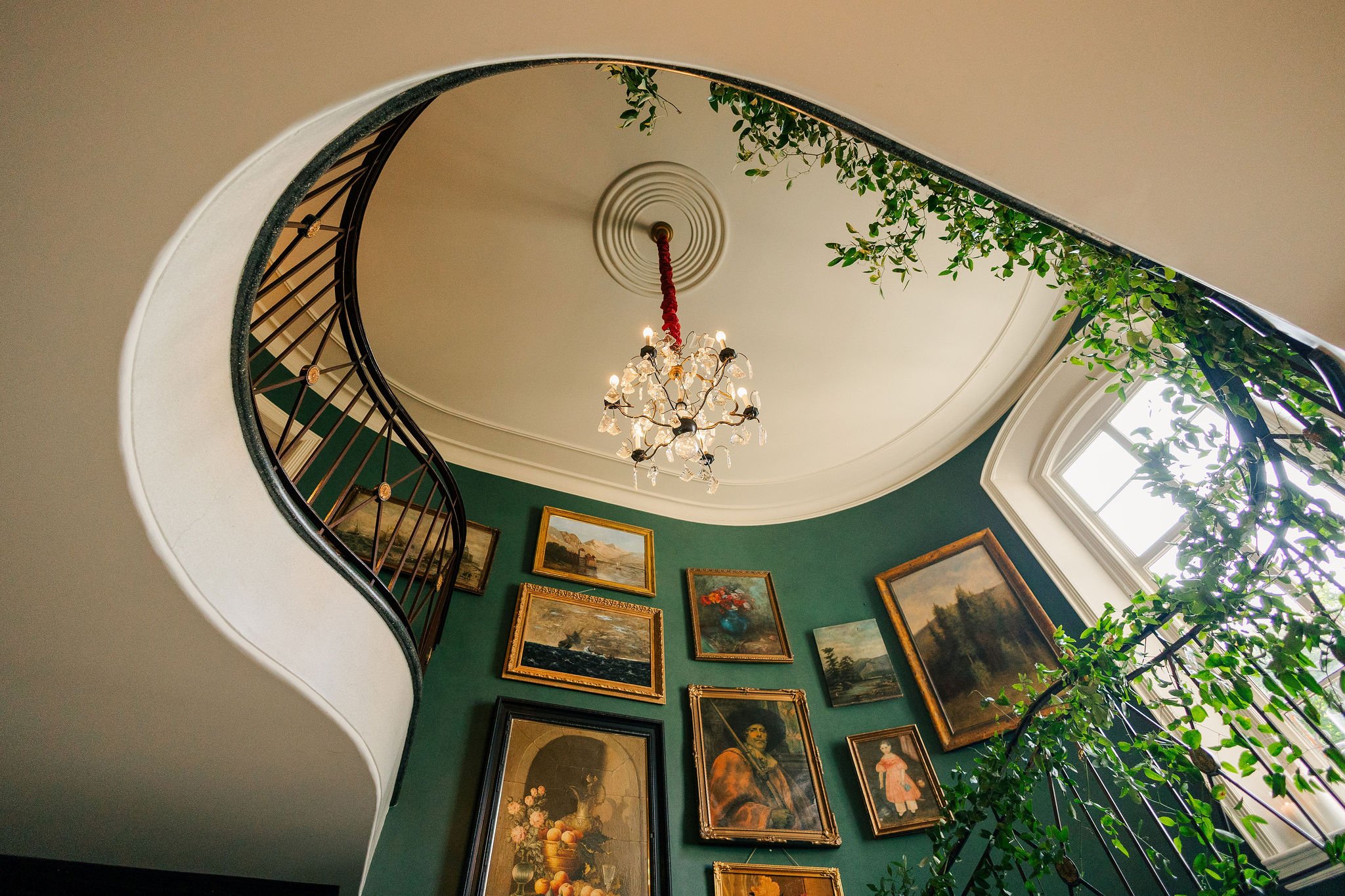 View of an elegant staircase with black railing, green walls decorated with framed paintings, a chandelier hanging from a ceiling, and a large window with abundant natural light.
