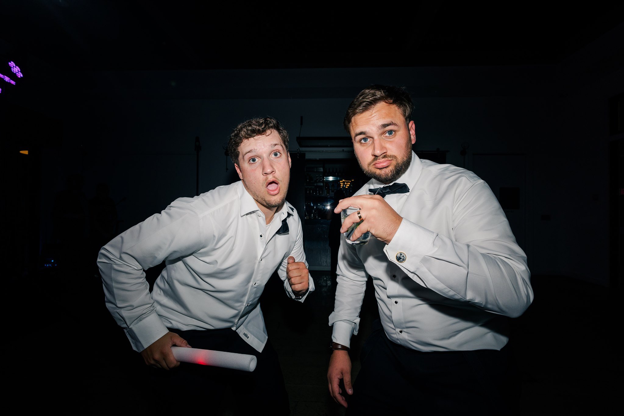 Two men in tuxedos making surprised and humorous faces at a party or event, standing close together in a dark room.