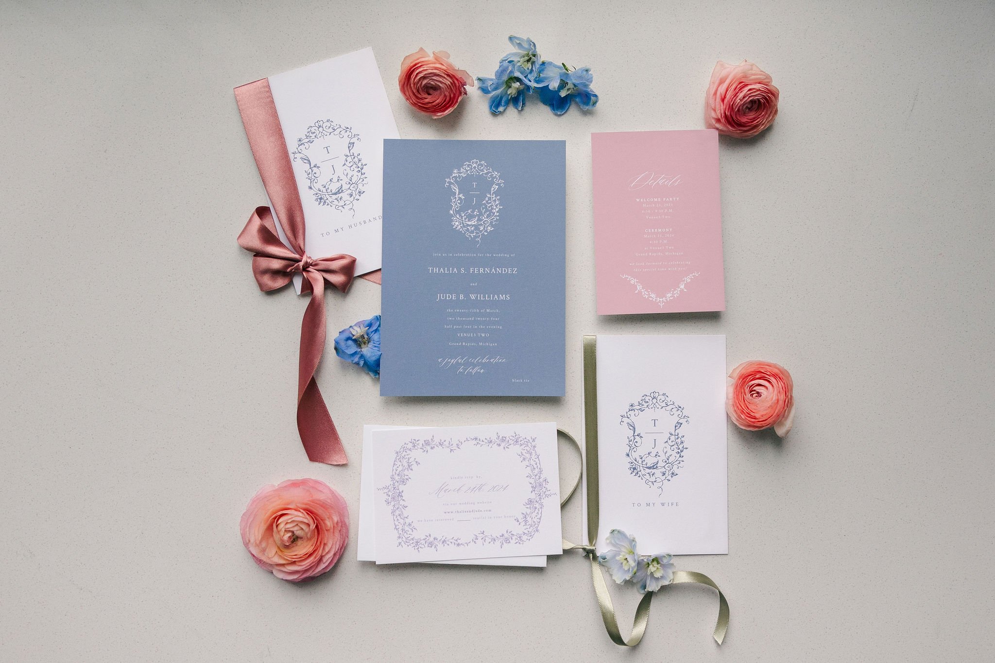 A flat lay of wedding invitation cards and floral decorations on a light surface. The cards feature elegant designs in pink, white, and blue, complemented by pink and blue flowers and satin ribbons.