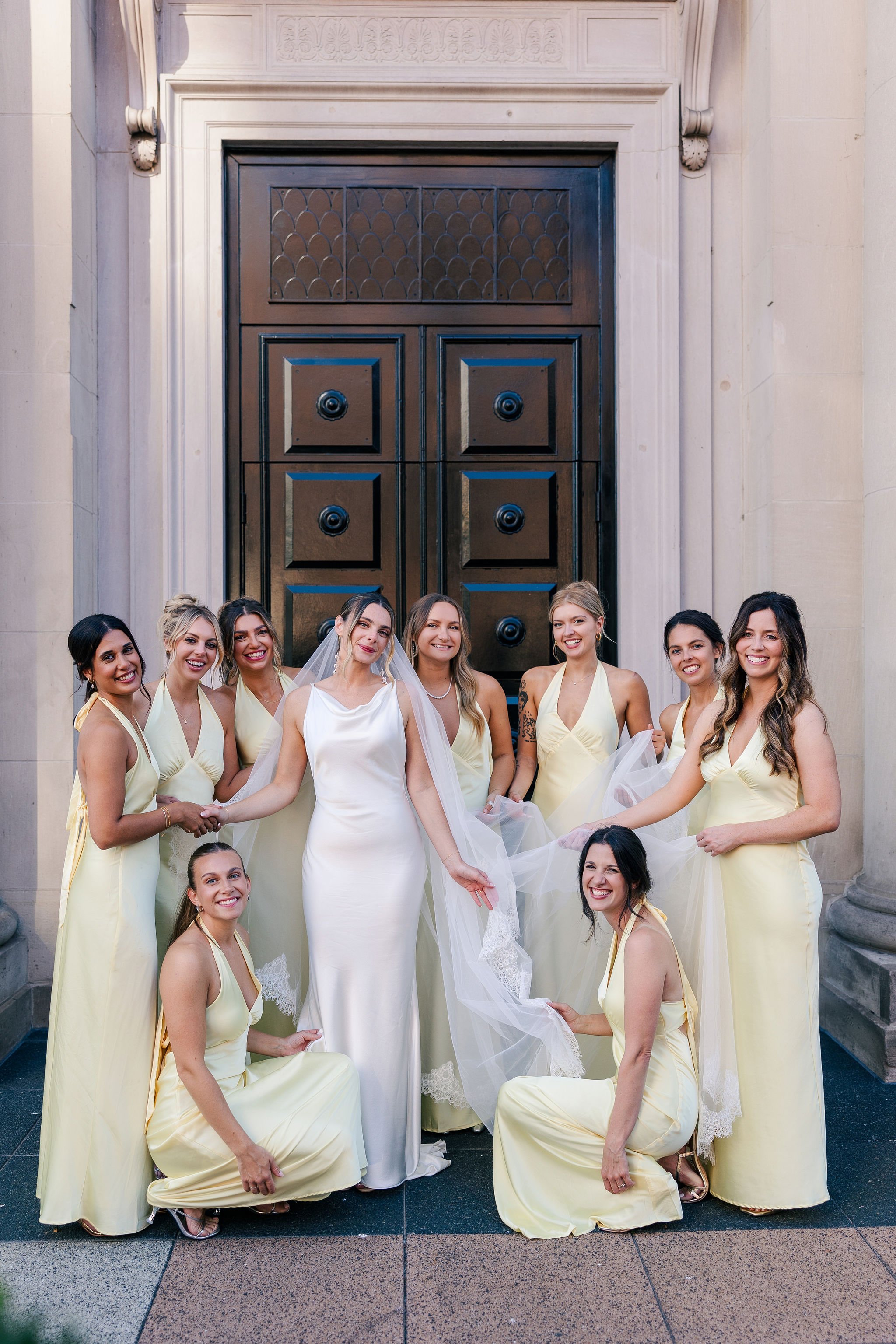 Bride and her nine bridesmaids in matching yellow dresses posing in front of a large black door at a wedding.