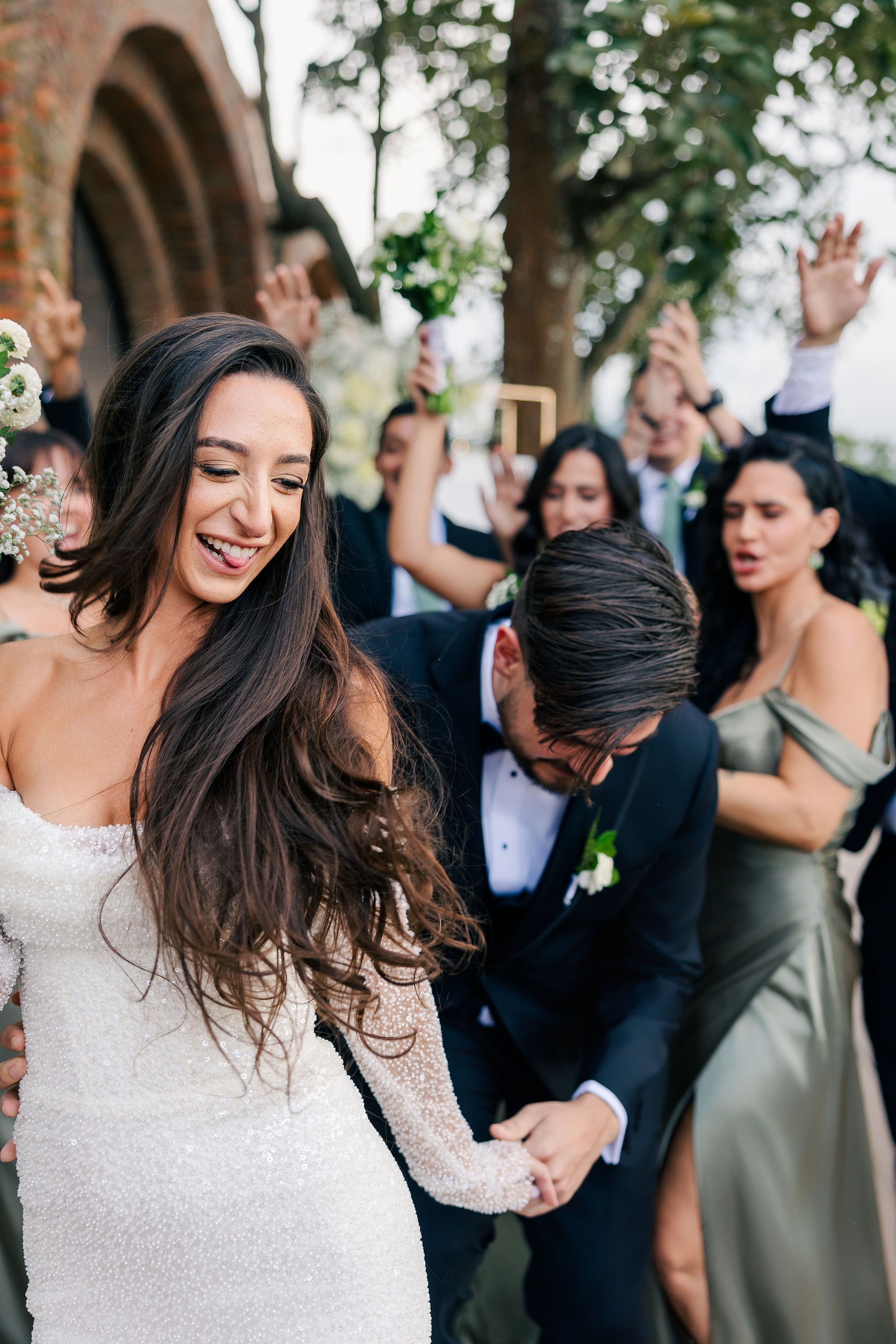 A bride and groom dancing at their wedding outdoors, surrounded by celebrating guests, some with raised hands and holding flowers, in front of a brick building with trees