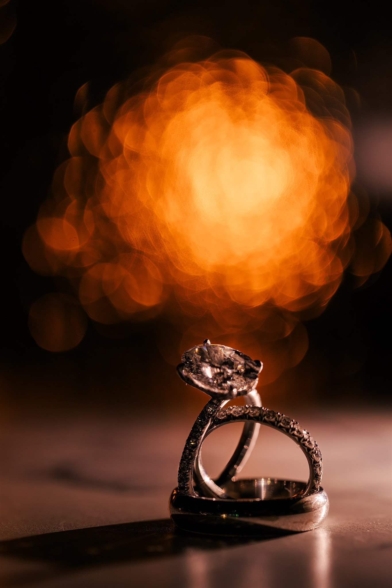 A close-up of two diamond engagement rings, one with a large central diamond and the other with small diamonds on the band, set against a dark background with warm, blurry lights in the distance.