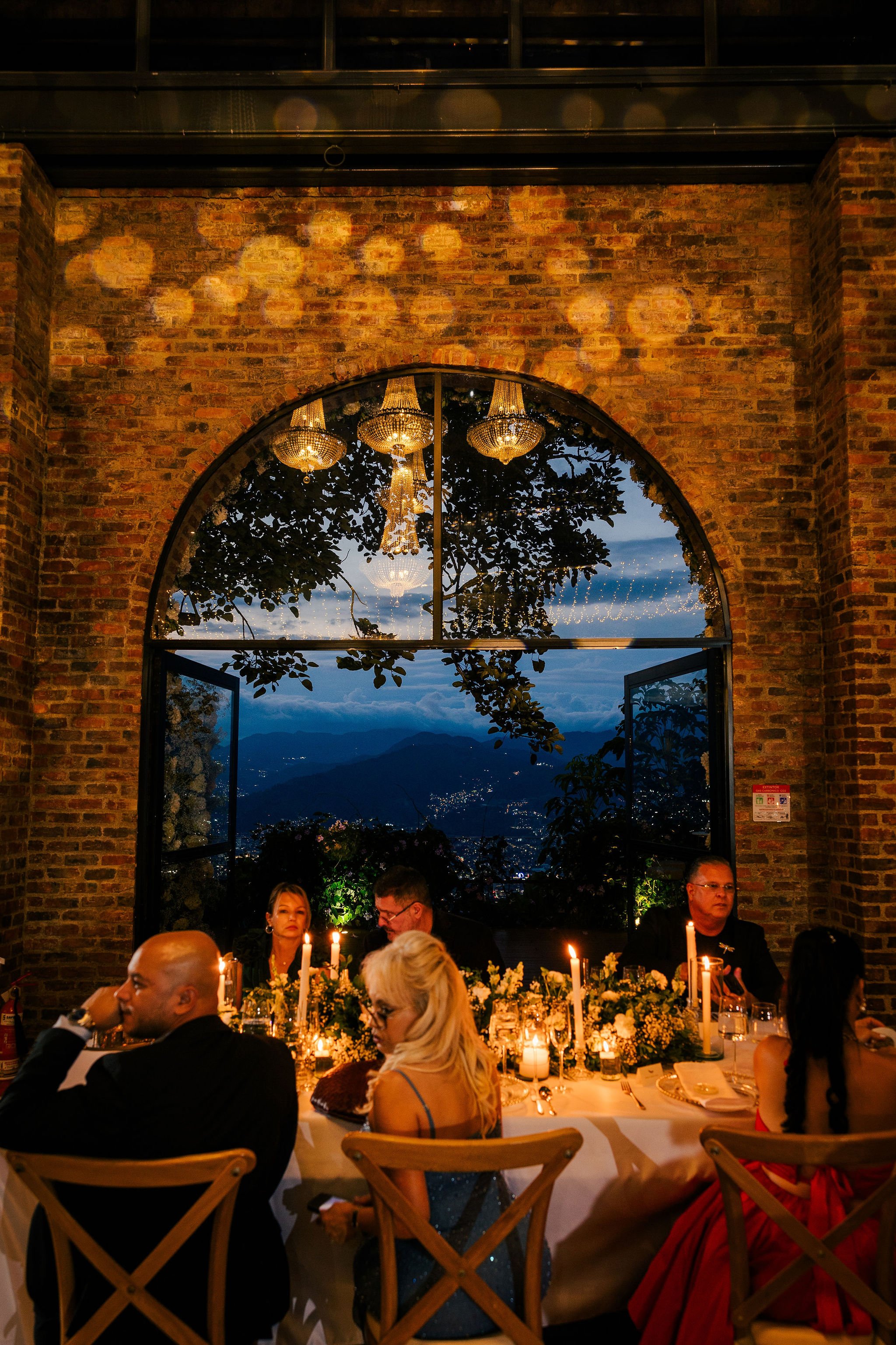 Indoor dining scene at night with a brick wall, large window revealing mountain landscape, and chandelier overhead. People seated at a decorated table with candles and flowers.