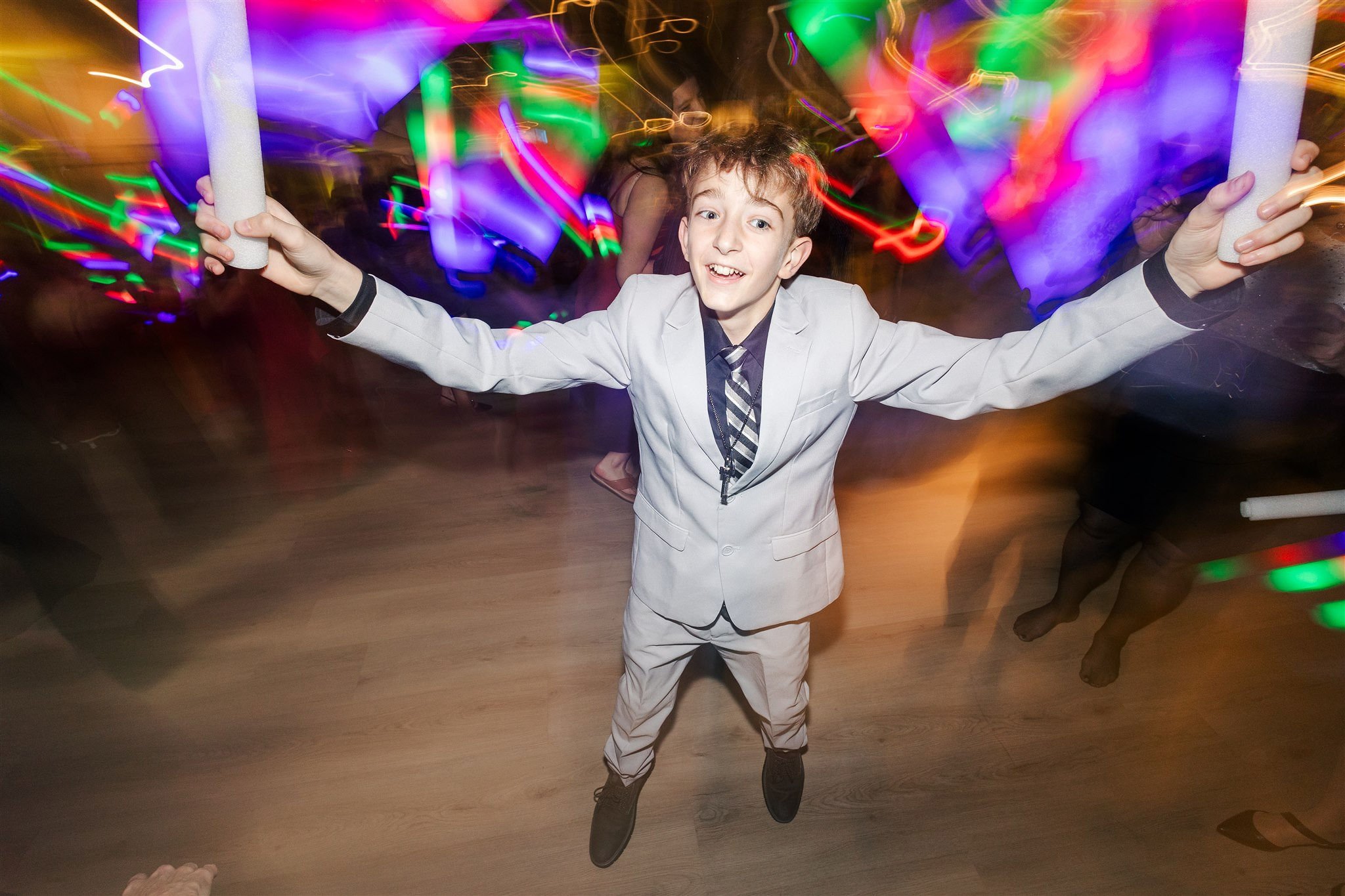 A young boy in a white suit with a tie, smiling with arms outstretched, at a lively party with colorful, blurred lights in the background.