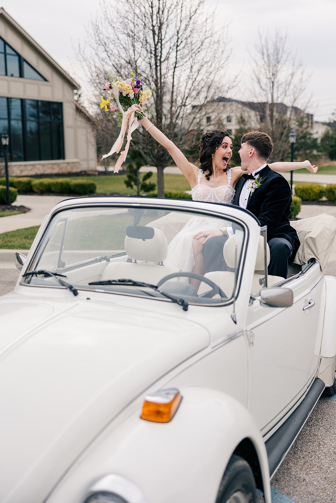 A newlywed couple celebrating in a vintage white convertible car, with the bride raising a bouquet of flowers and both smiling in an outdoor setting.