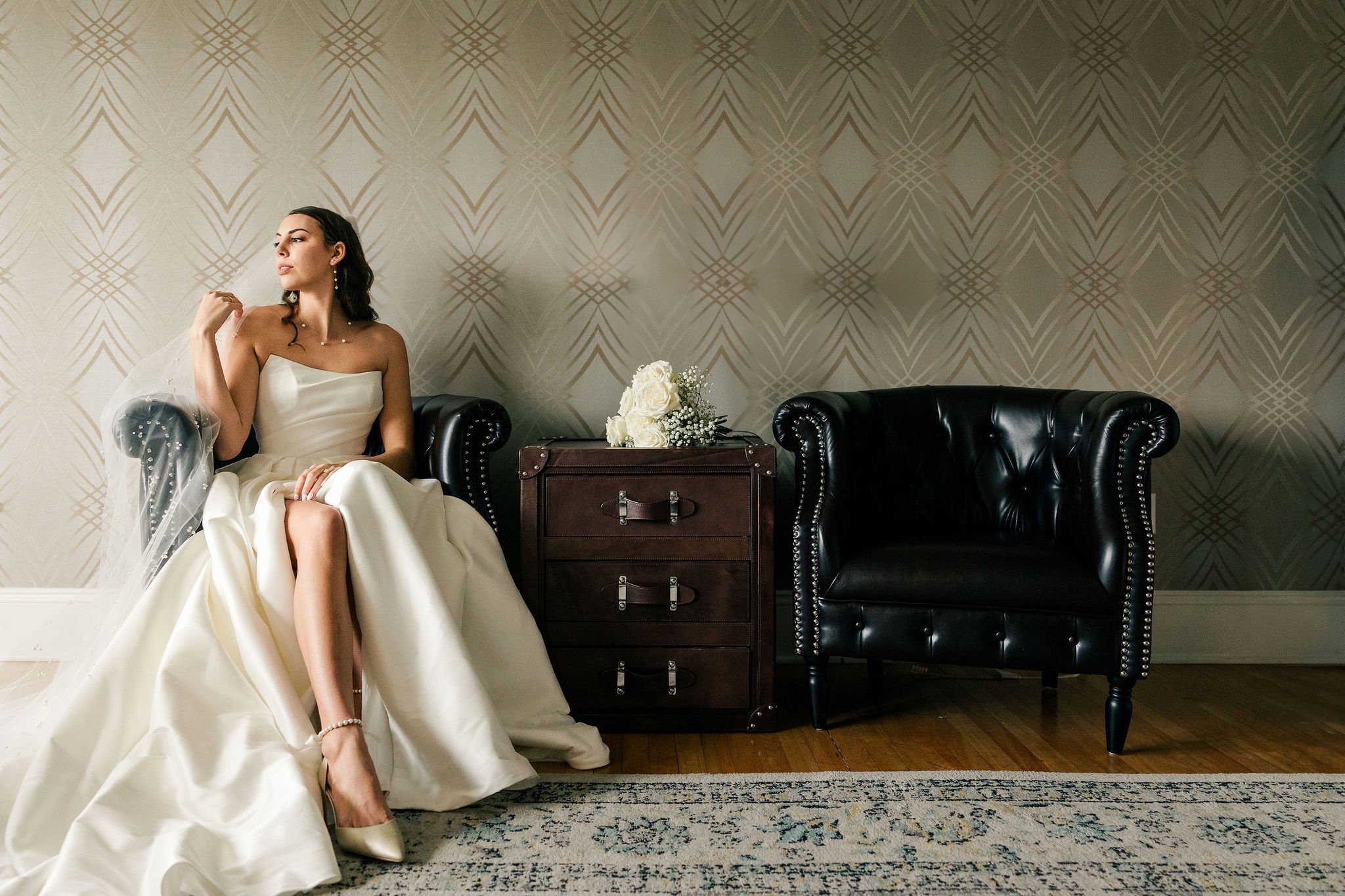 A woman in a white wedding dress and veil sitting on a black leather armchair next to an empty armchair and a small wooden table with white roses, against a patterned wall.