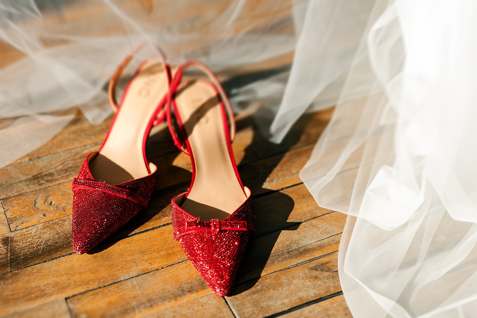 Red glittery high-heeled shoes with bows on a wooden floor, partially covered by a sheer white fabric.
