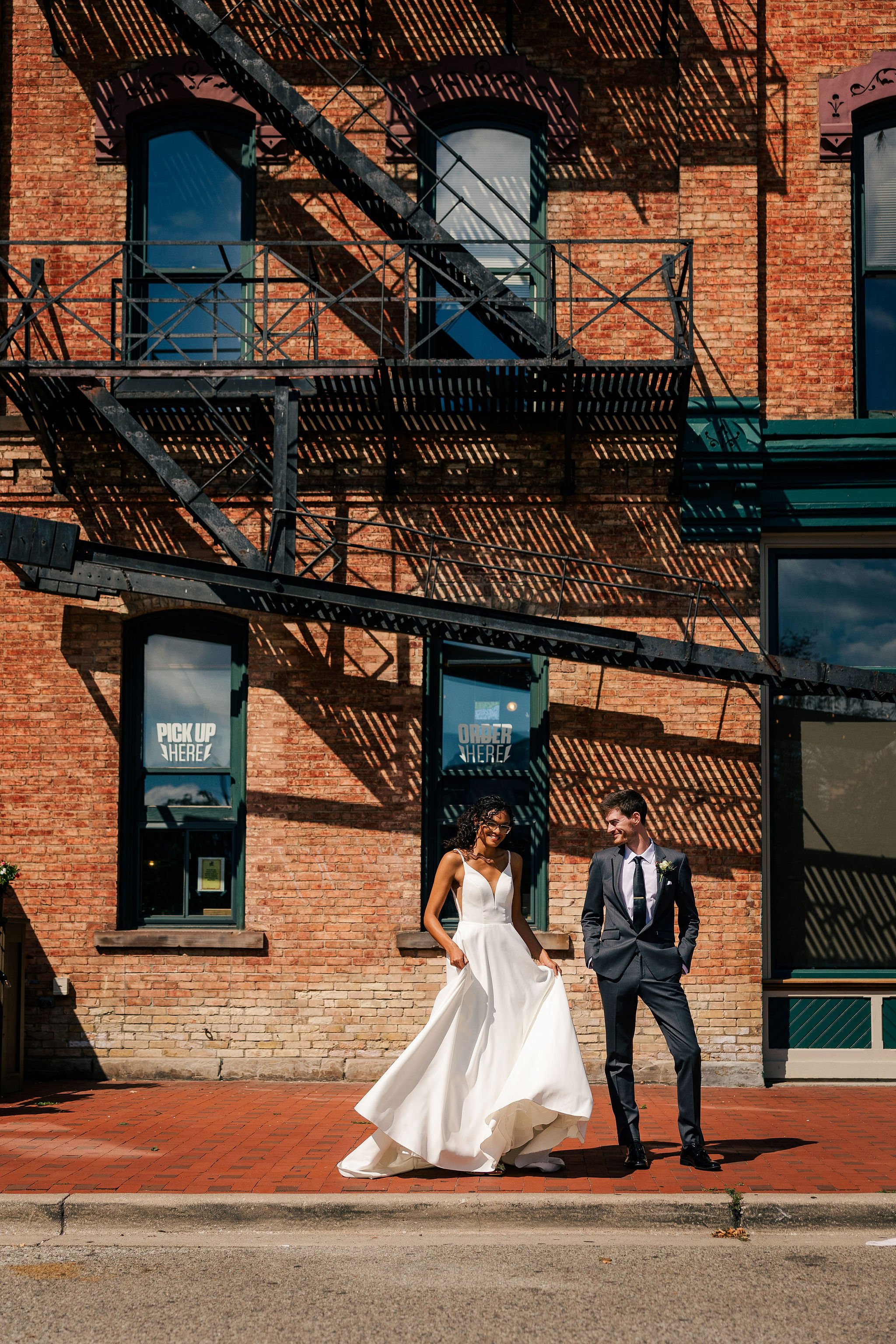 A bride in a white gown and a groom in a black suit standing on a brick sidewalk outside a brick building with fire escapes. The bride is smiling and holding her dress, while the groom is looking at her, both appearing happy.