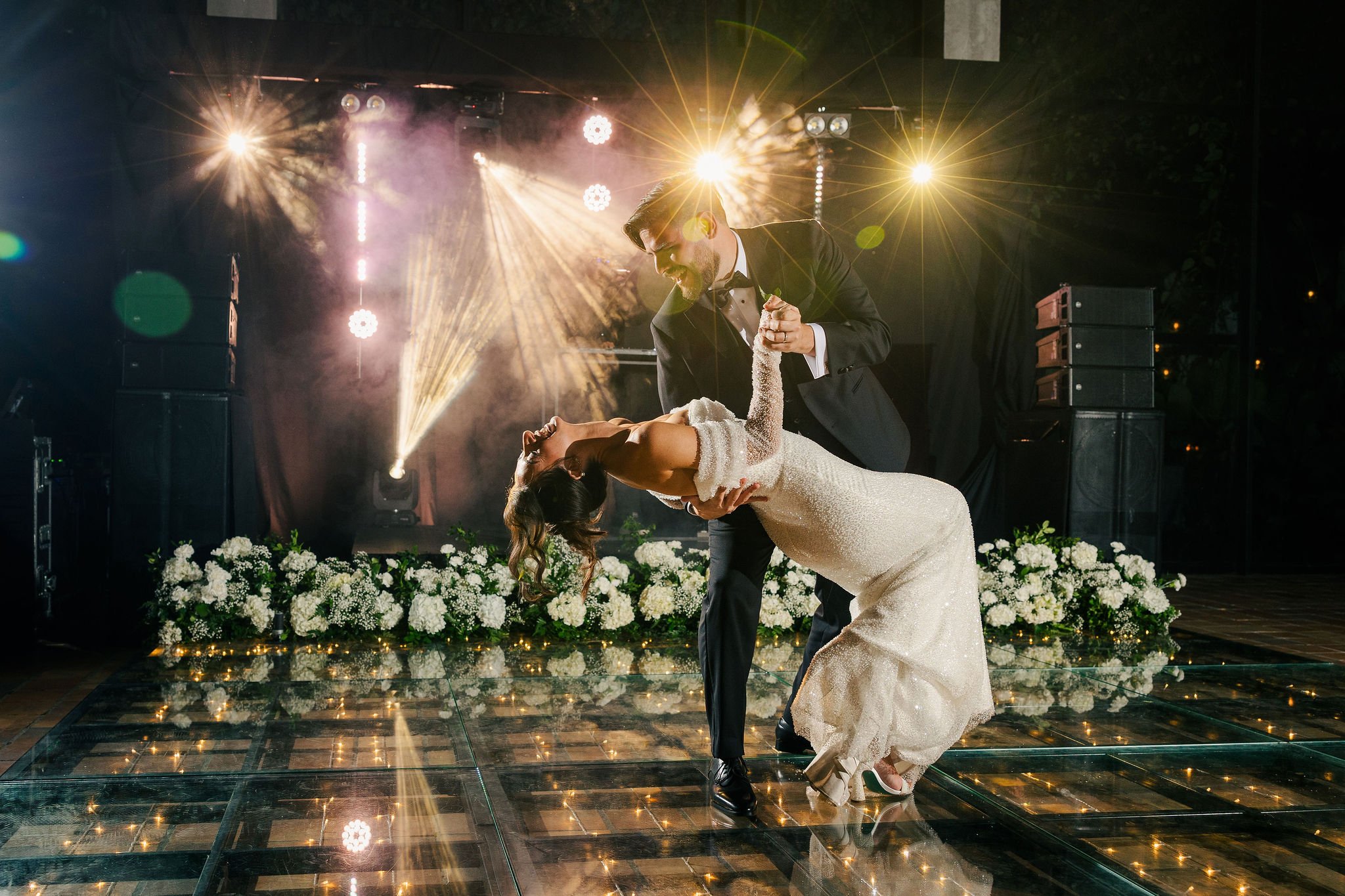 A couple dancing at their wedding reception with dramatic lighting, a floral backdrop, and a dark dance floor.