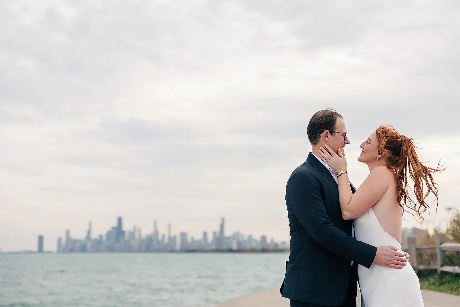 A man and woman in wedding attire embracing outdoors near water with a city skyline in the background.