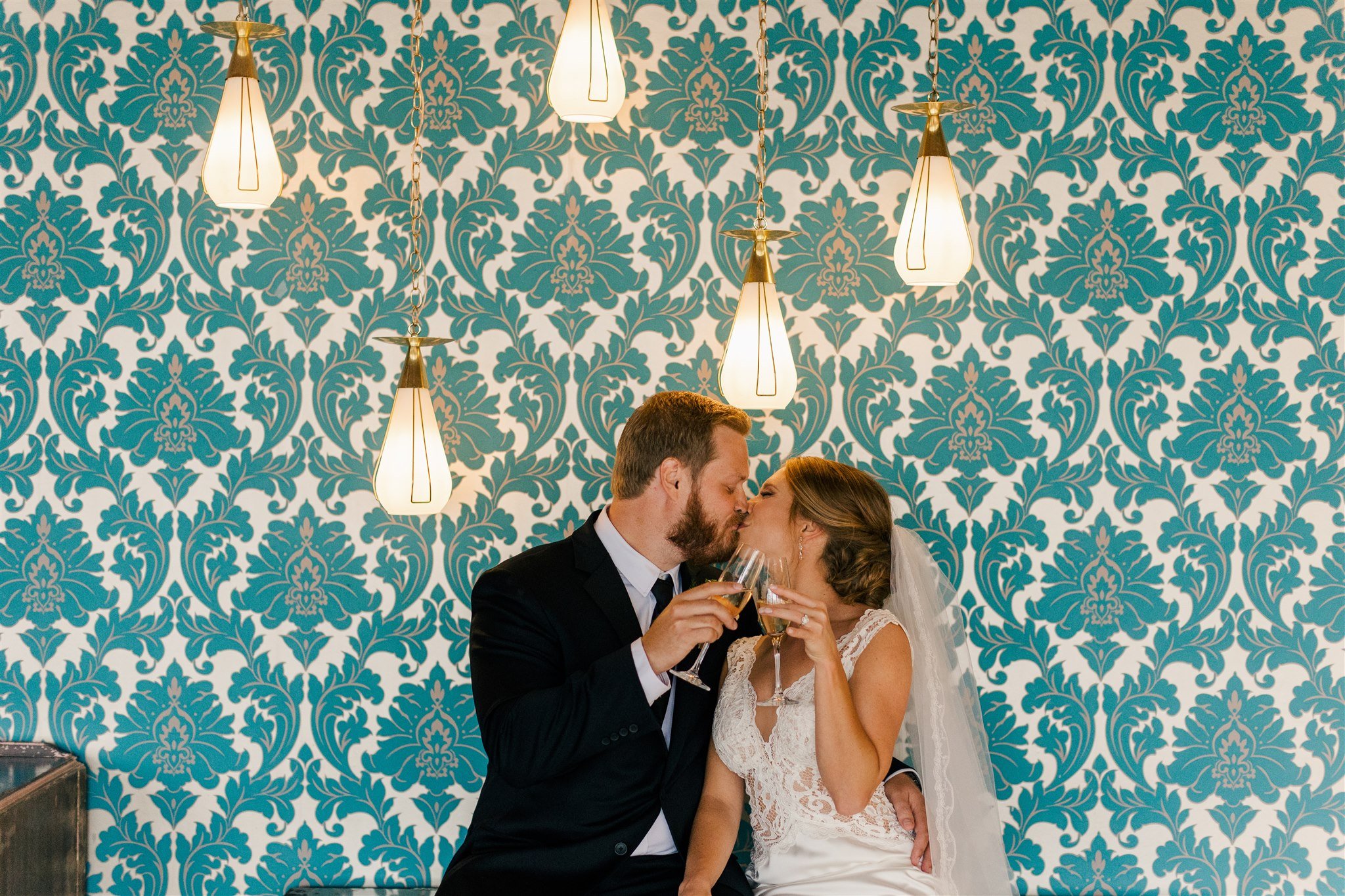 Bride and groom in wedding attire sharing a kiss and holding glasses of champagne in front of a teal damask wallpaper with hanging pendant lights.